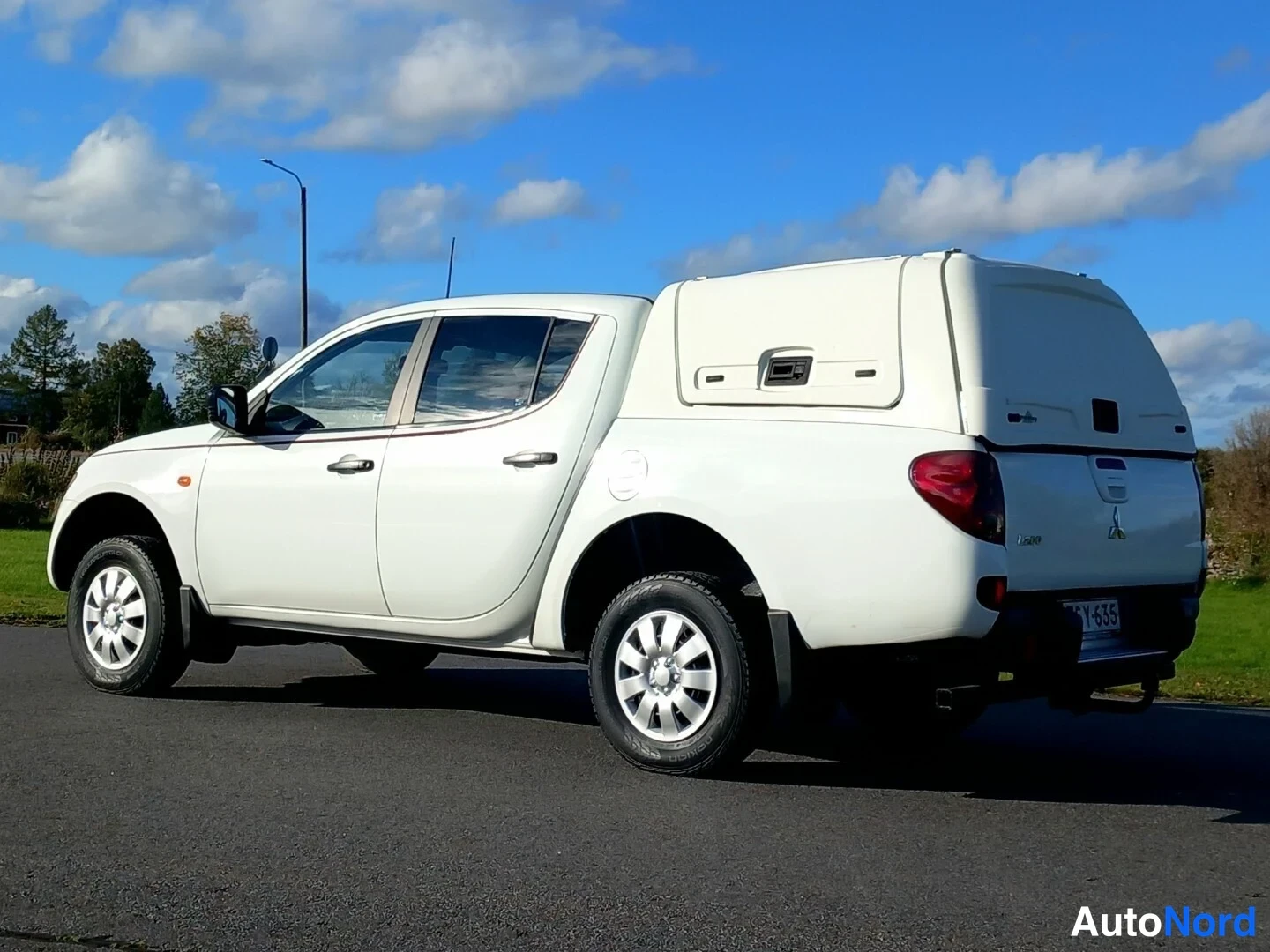 Takaviisto: Valkoinen Mitsubishi L200 takaviistosta oikealta, rekisteritunnus, neljä ovea ja lisäkori lavalla.