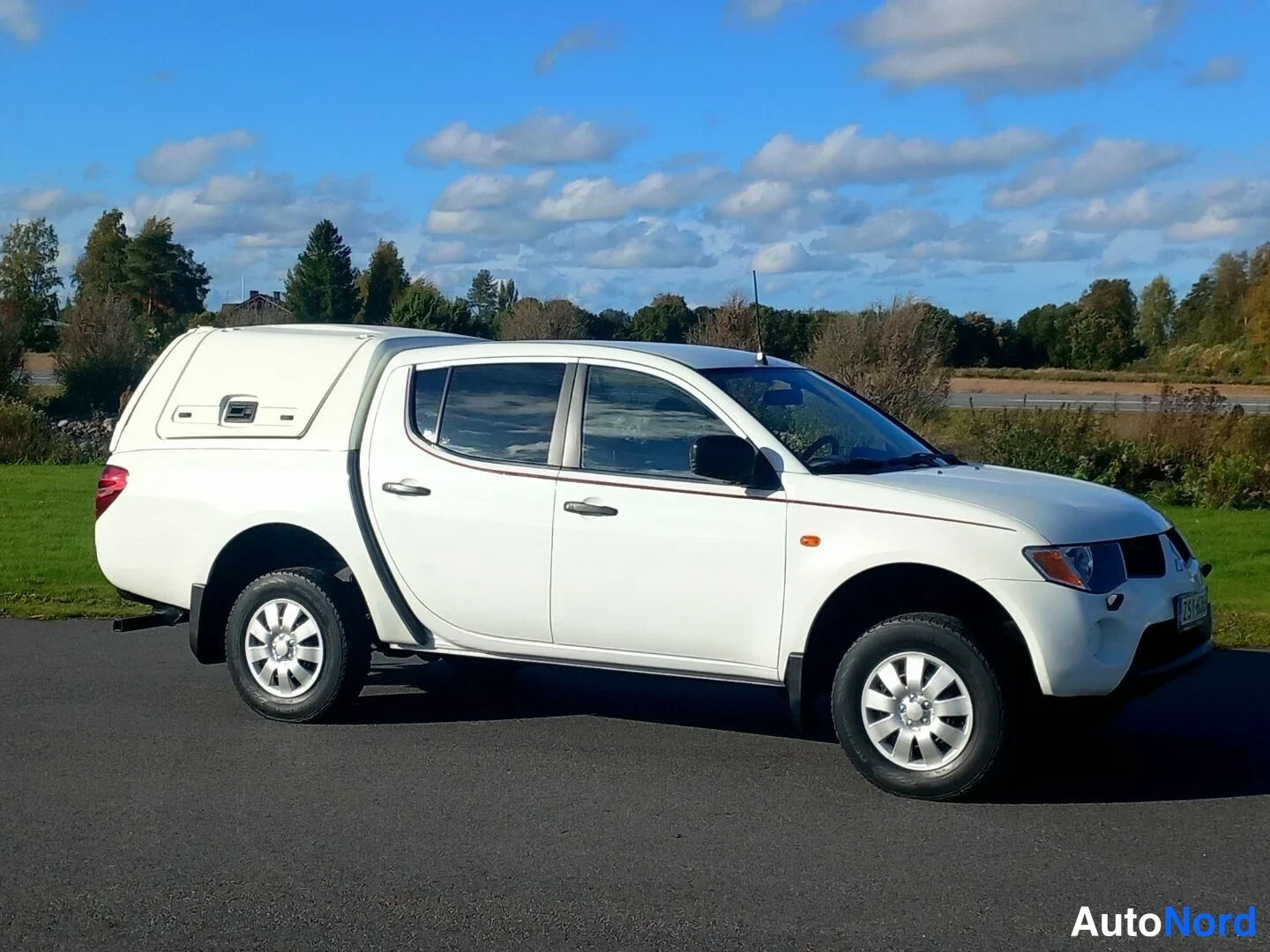 Etuviisto, nokka oikealle: Valkoinen Mitsubishi L200 -avolava kylki- ja keulakulma oikealta, hardtop lavalla, tausta sininen taivas ja pellot.