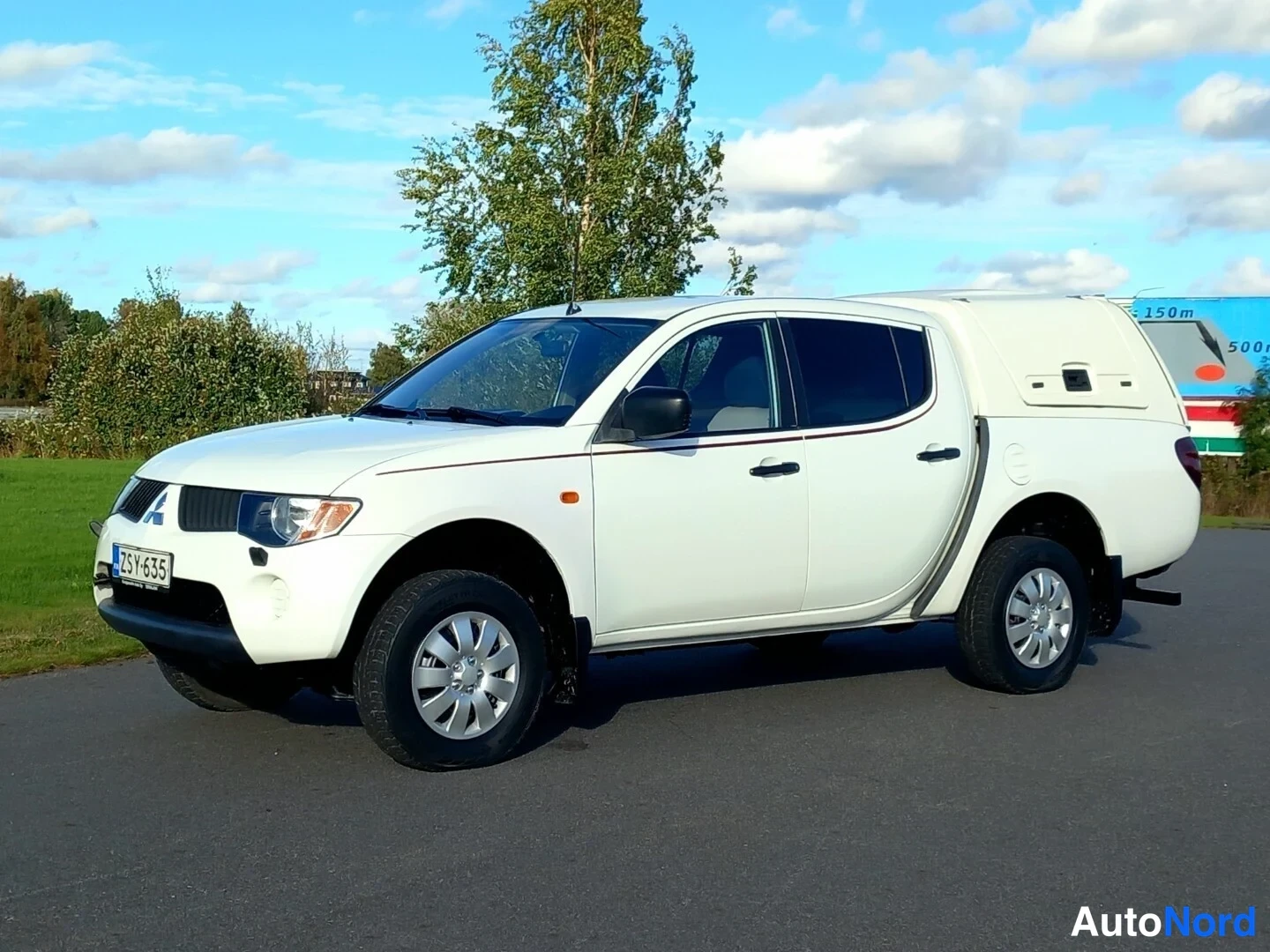 Etuviisto, nokka vasemmalle: Valkoinen Mitsubishin avolava-auto sivulta edestä kuvattuna, neliovinen ohjaamo ja kiinteä kupe suojana lavalla.