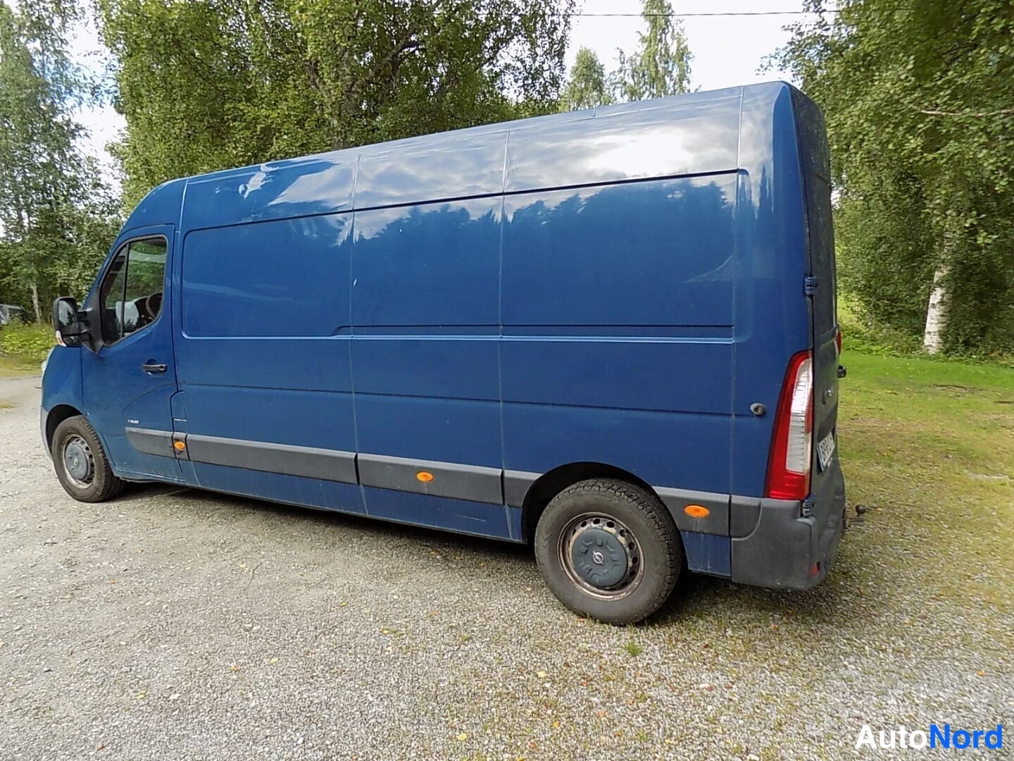 Takaviisto: Tummansininen pakettiauto takaviistosta oikealta sorapohjaisella paikalla, takaosassa punaiset takavalot.
