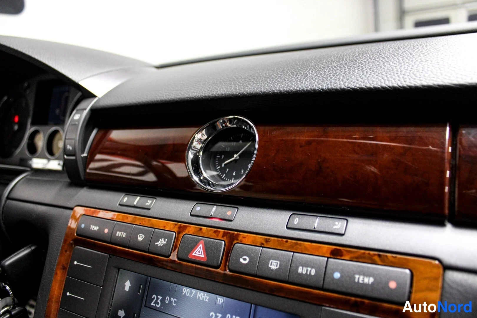 Muu: Kojelaudan puupaneloitu alue ja kromireunainen analoginen kello; alla ilmastointipainikkeet TEMP, AUTO ja varoituskolmio, näytössä 23,0 °C ja 90.7 MHz.