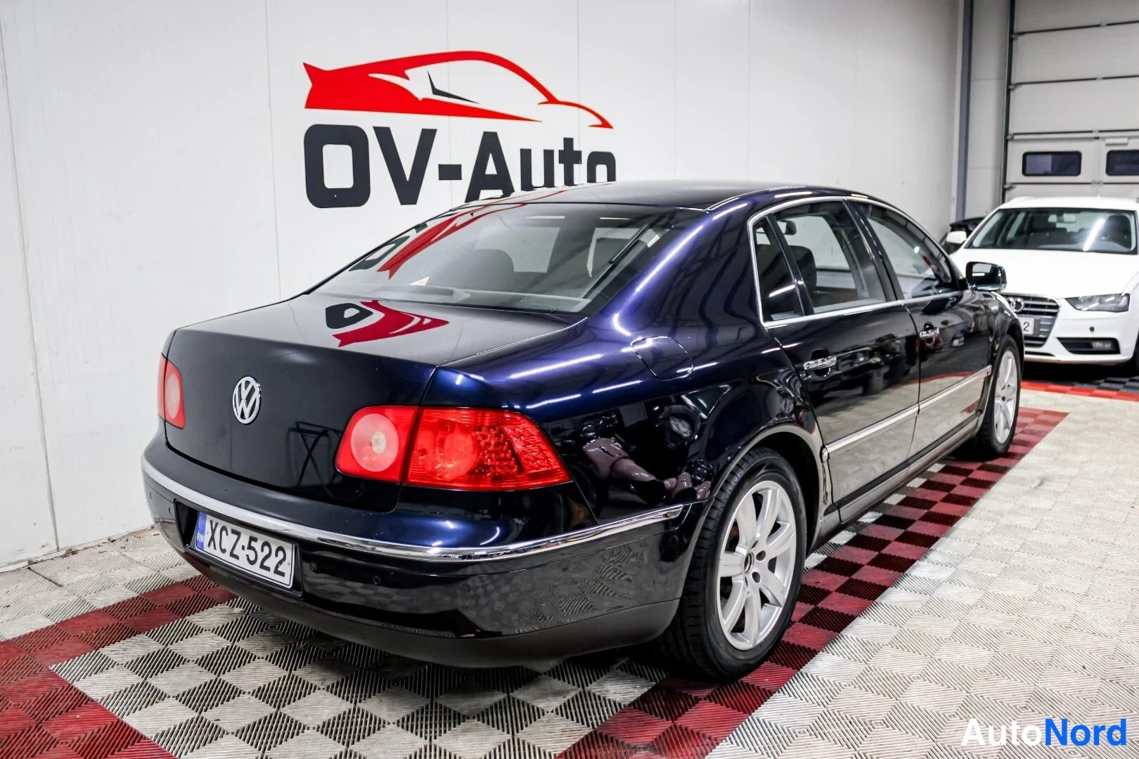 Takaviisto: Sinisen musta Volkswagen Phaeton -sedan takaa sivulta kuvattuna hallissa, rekisterikilpi.