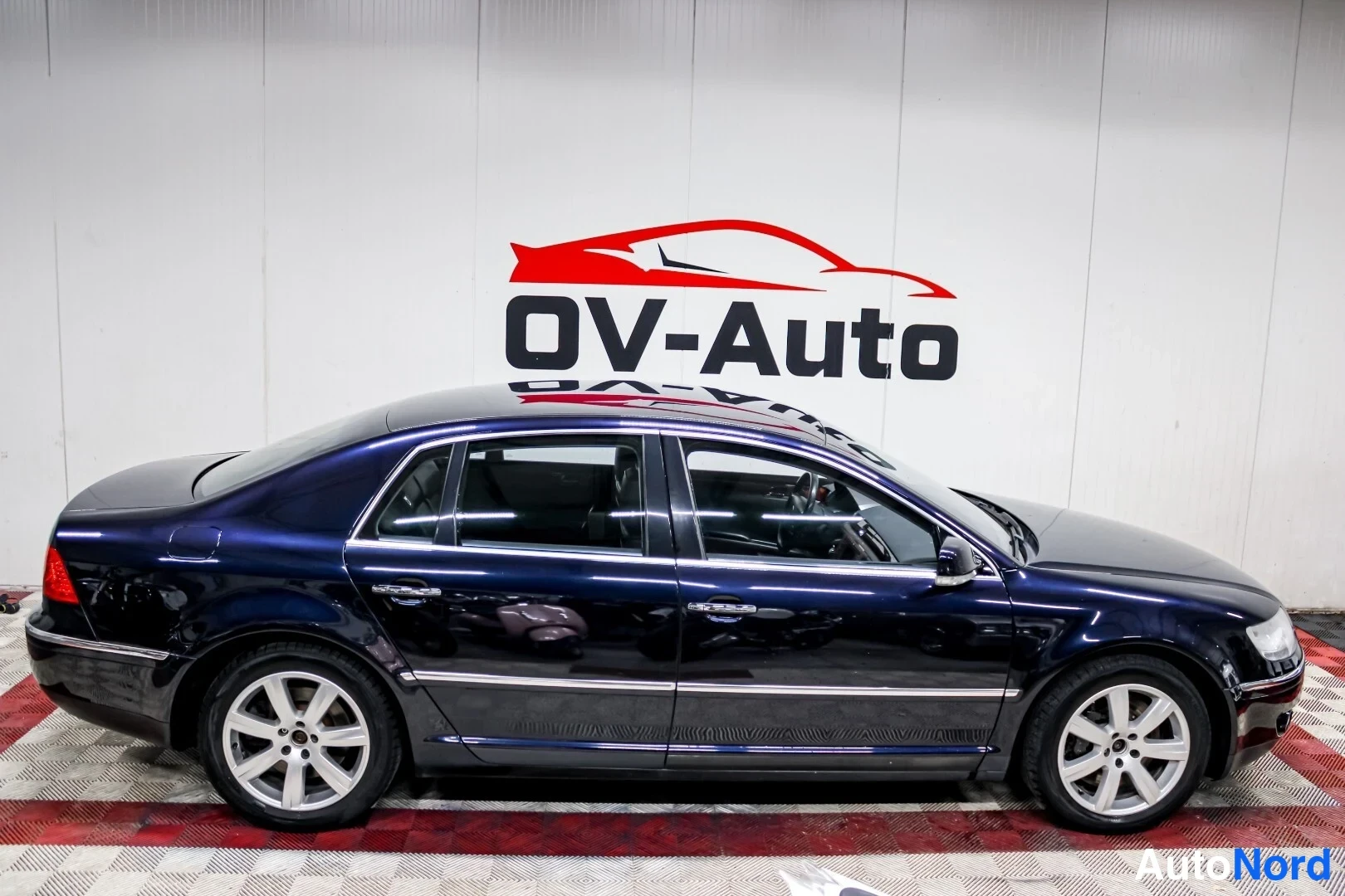 Sivulta: Tummansininen Volkswagen Phaeton -sedan sivuprofiilissa hallissa, hopeiset peltivanteet ja kromilista ovessa.