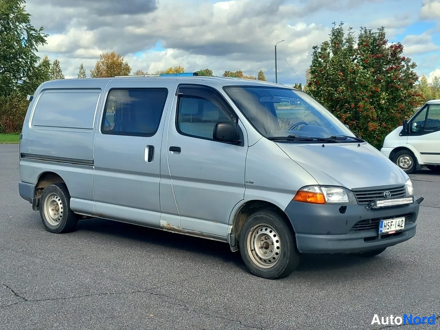 Etuviisto, nokka oikealle: Toyota Hiace -pakettiauto hopeanvärisenä sivukulmasta, keula oikealla, rekisterikilpi.