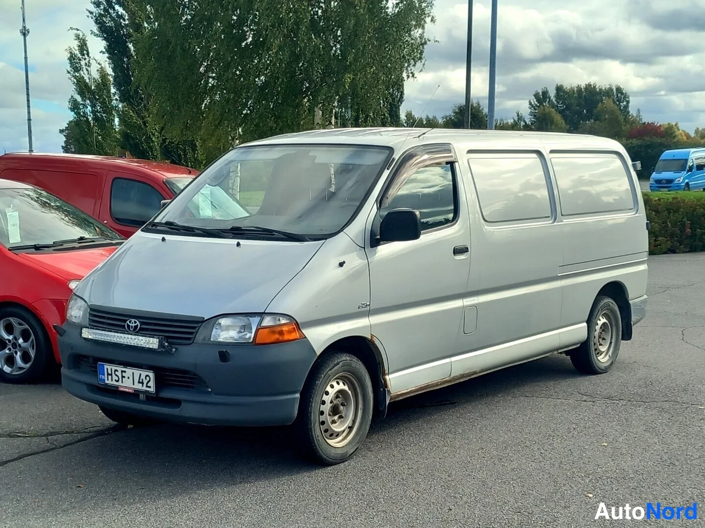 Etuviisto, nokka vasemmalle: Toyota Hiace -pakettiauto hopeanvärisenä etukulmasta vasemmalta, rekisterikilpi.
