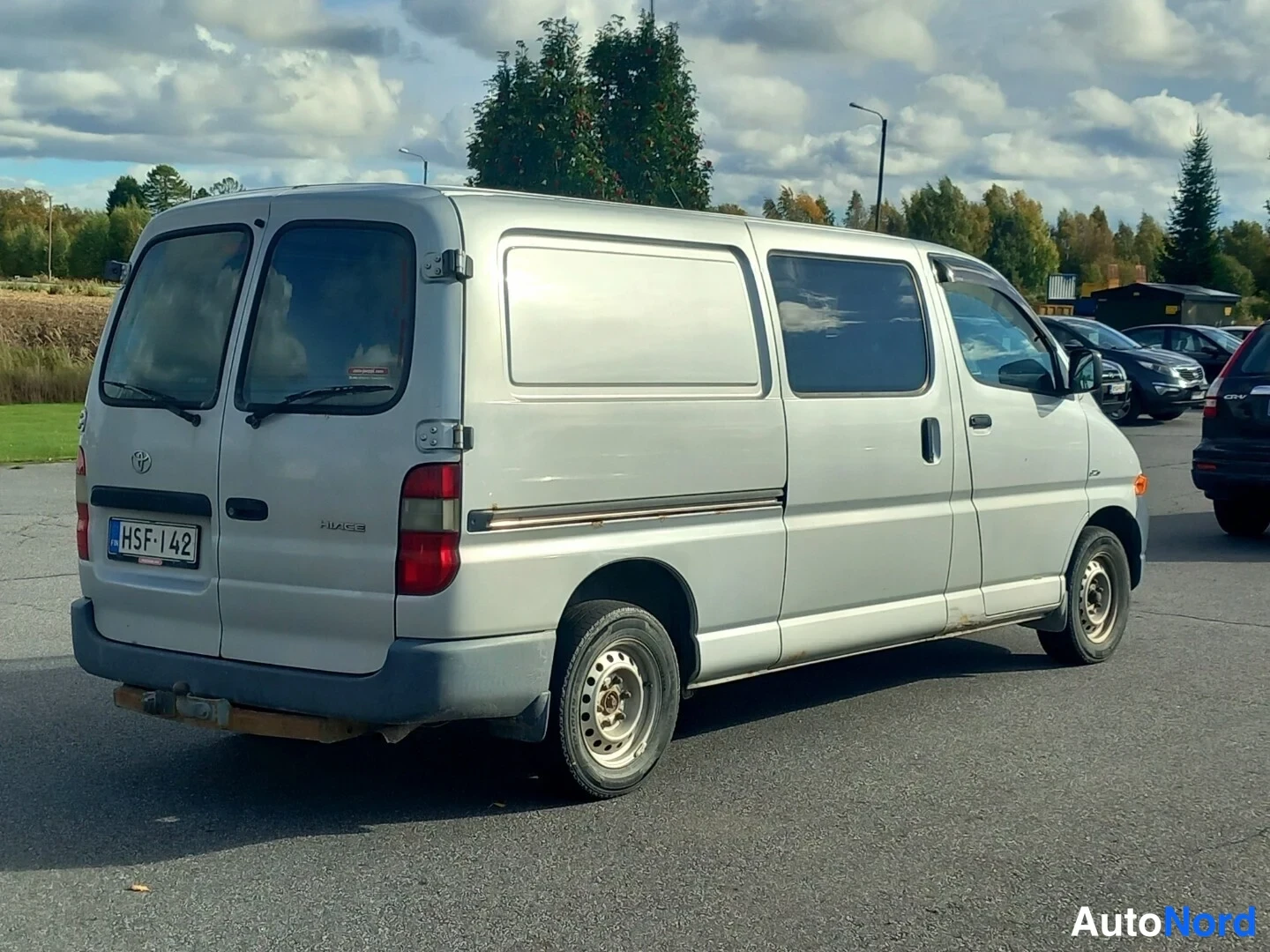 Takaviisto: Toyota Hiace -pakettiauto takaviistosta, hopeisenä; rekisterikilpi, takaluukun Toyota-merkki ja Hiace-teksti näkyvät.