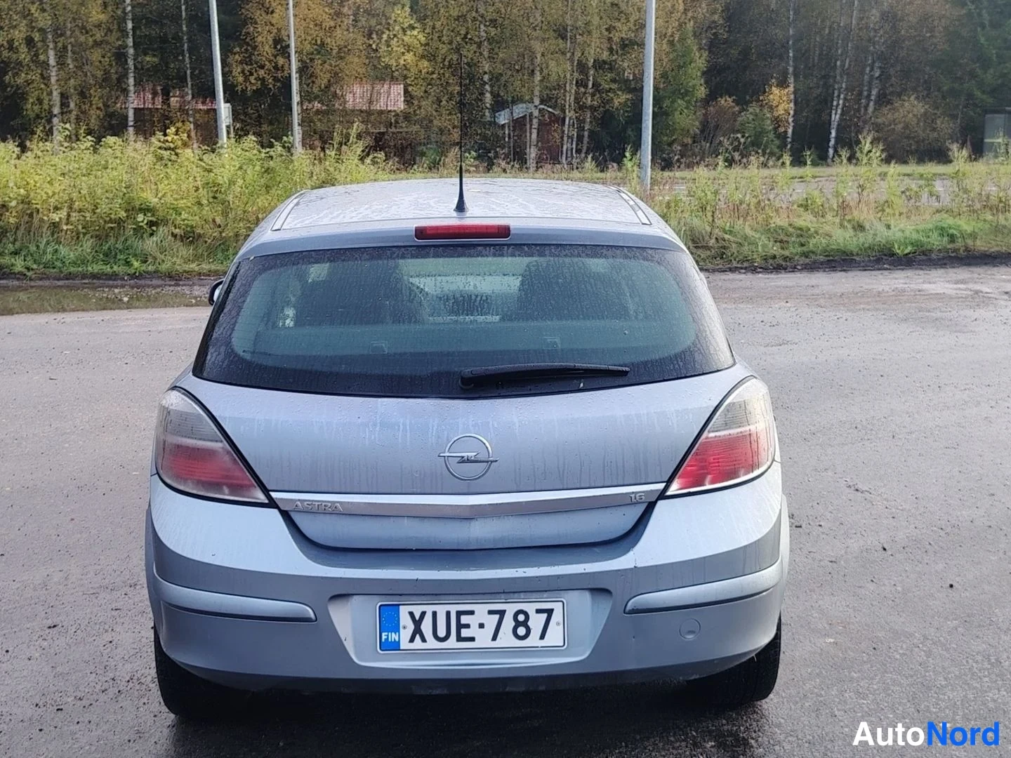 Takaa: Auton takaosa suoraan takaa: hopeanharmaa Opel Astra, rekisterikilpi, takavalot ja Opel-merkki näkyvissä, märkä asfaltti ja puita taustalla.