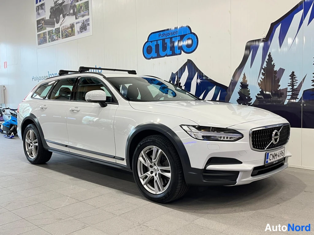 Etuviisto, nokka vasemmalle: Etu- ja oikea puoli kolmannes näkymä valkoisesta Volvo V90 Cross Country -henkilöautosta, jossa on katonrakelaitteet ja kevytmetallivanteet.
