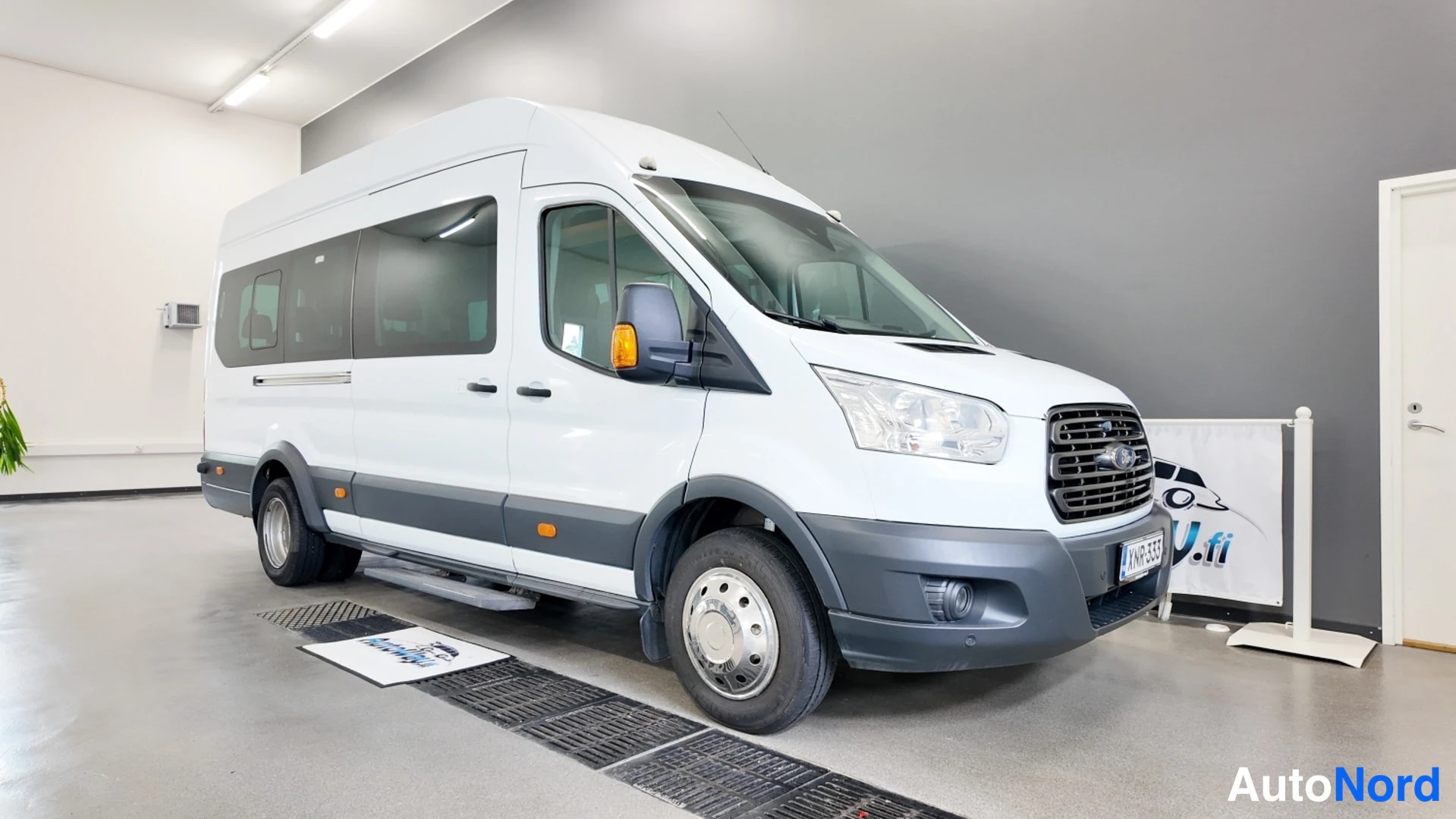 Etuviisto, nokka oikealle: Valkoinen Ford Transit -pakettiauto kulmassa kuvattuna sisätiloissa, etuosa ja sivu näkyvissä.