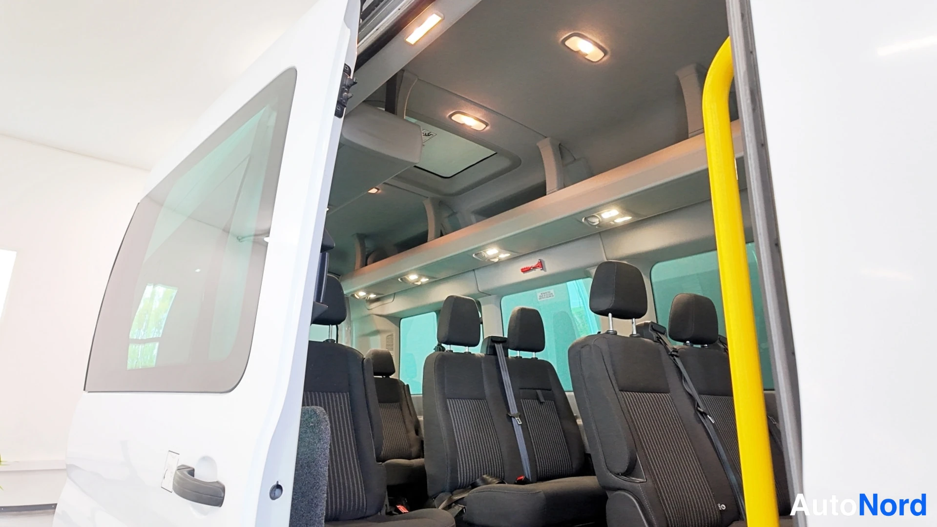 Muu: Ford Transitin sisätilat takaovasta kuvattuna: tummat penkit ja keltainen tukikahva.