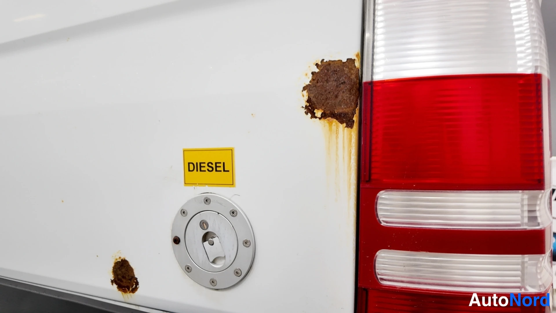 Yksityiskohta: Valkoisen auton takakulman lähikuva: punainen takavalaisin,Diesel-täyttöaukko ja selvää pintaruostetta.