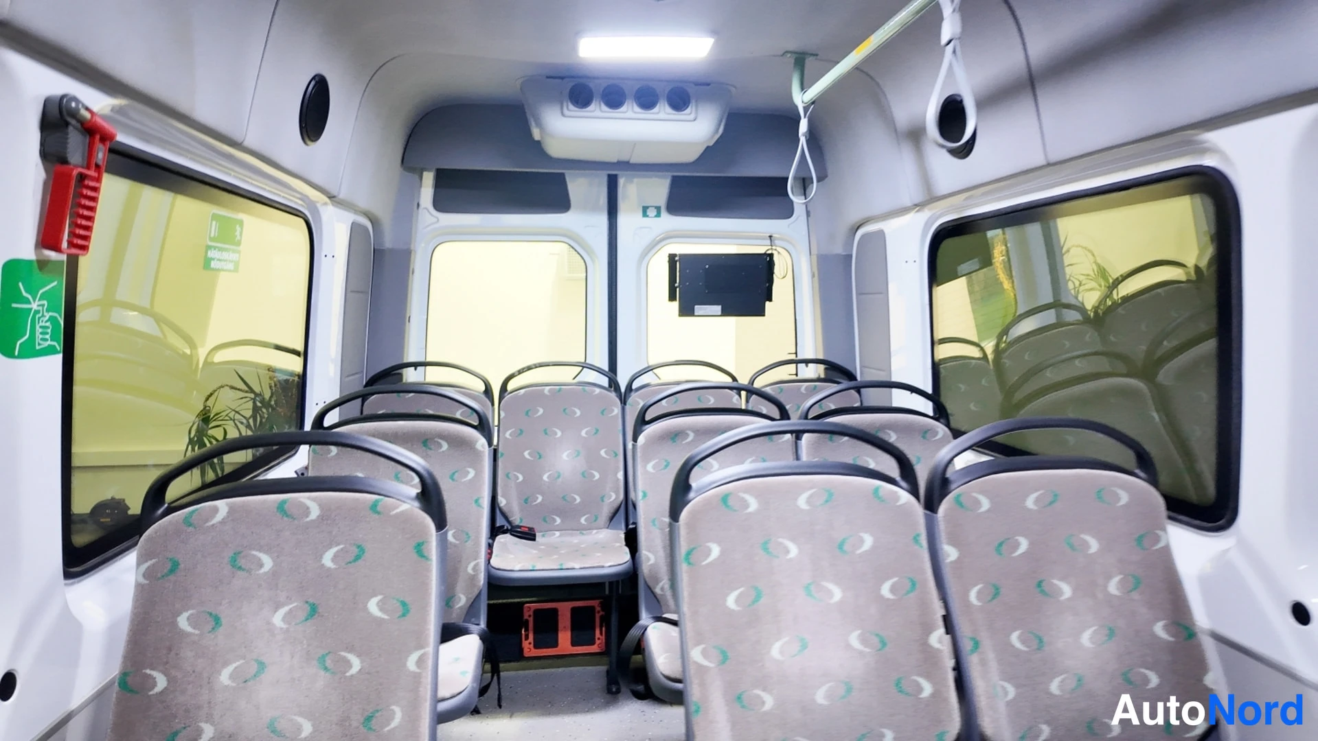 Muu: Pakettibussin matkustamoon takaa päin: kaksi riviä kudottupäällysteisiä istuimia, katon LED-valo ja hoidinkahvat.