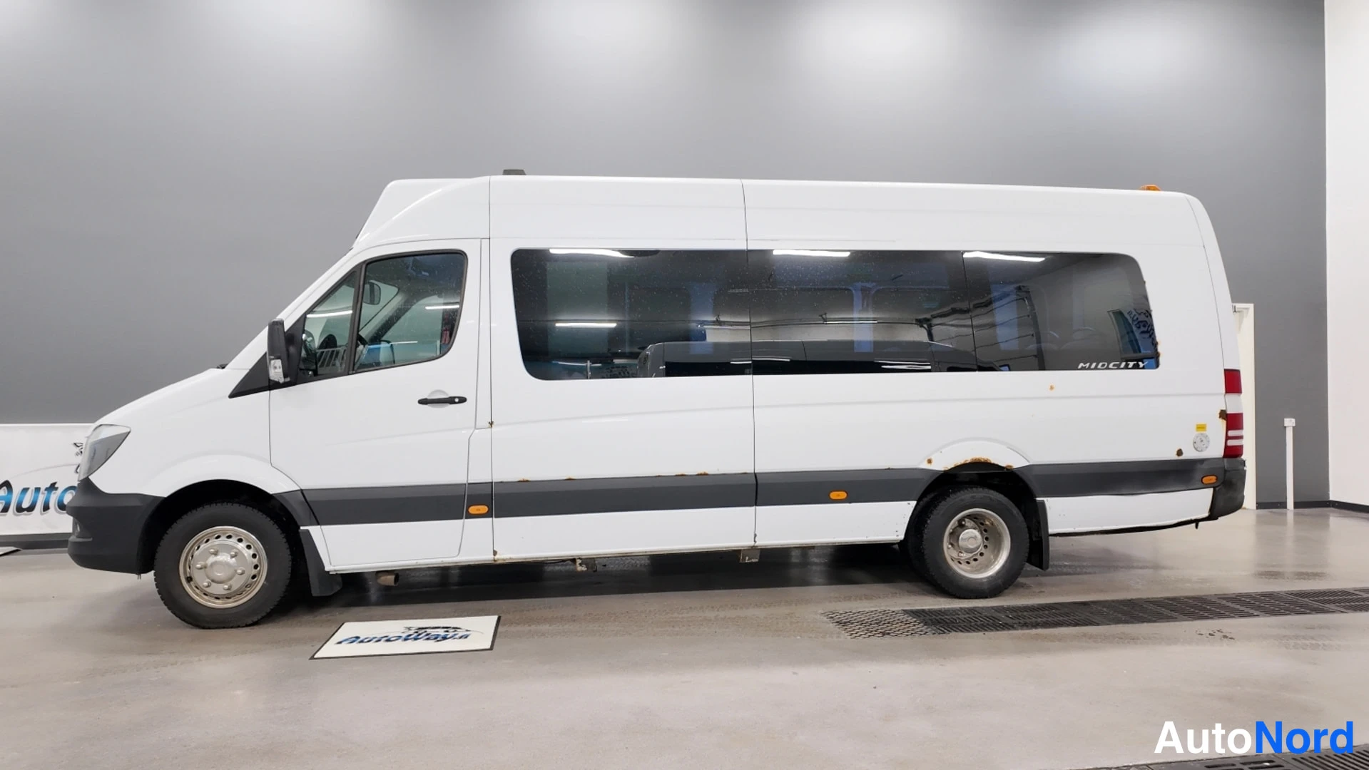 Sivulta: Mercedes-Benz Sprinter -pakettiauto sivukuva sisätiloissa, koko vasen kylki näkyvissä, valkoinen koripeli ja tummat ikkunat.
