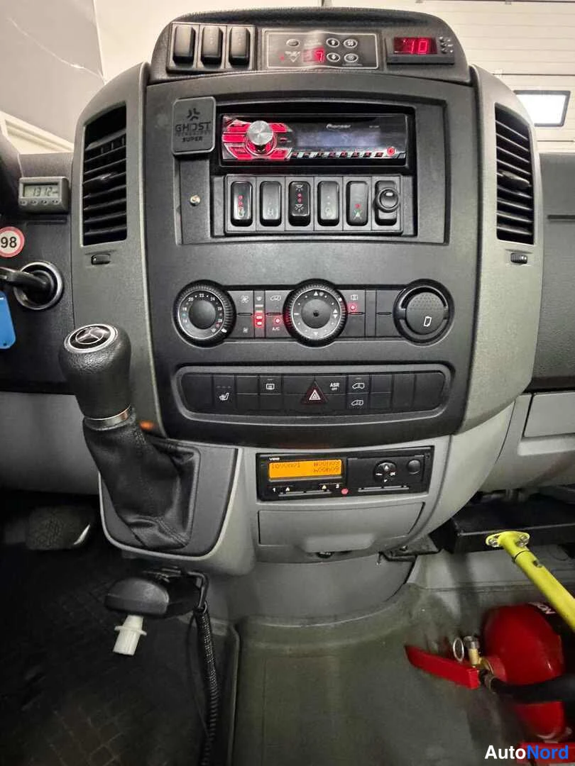 Edestä: Mercedes-Benz Sprinterin kojelaudan keskiö: Pioneer-radio, ohjauskytkimet, lämpötilanappulat, varoituskolmio, vaihtajan keppi ja palosammutin lattialla.