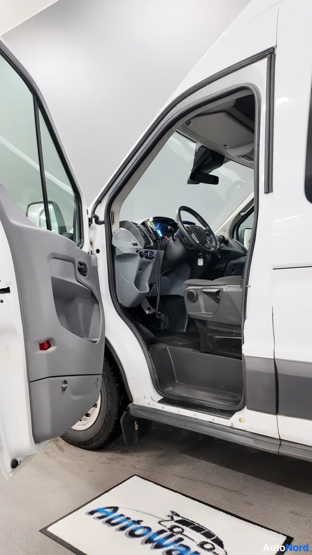 Sivulta: Pakettiauton kuljettajan ovi on auki ja ohjaamo näkyy: ohjauspyörä, kojetaulu ja istuin. Auto on valkoinen.
