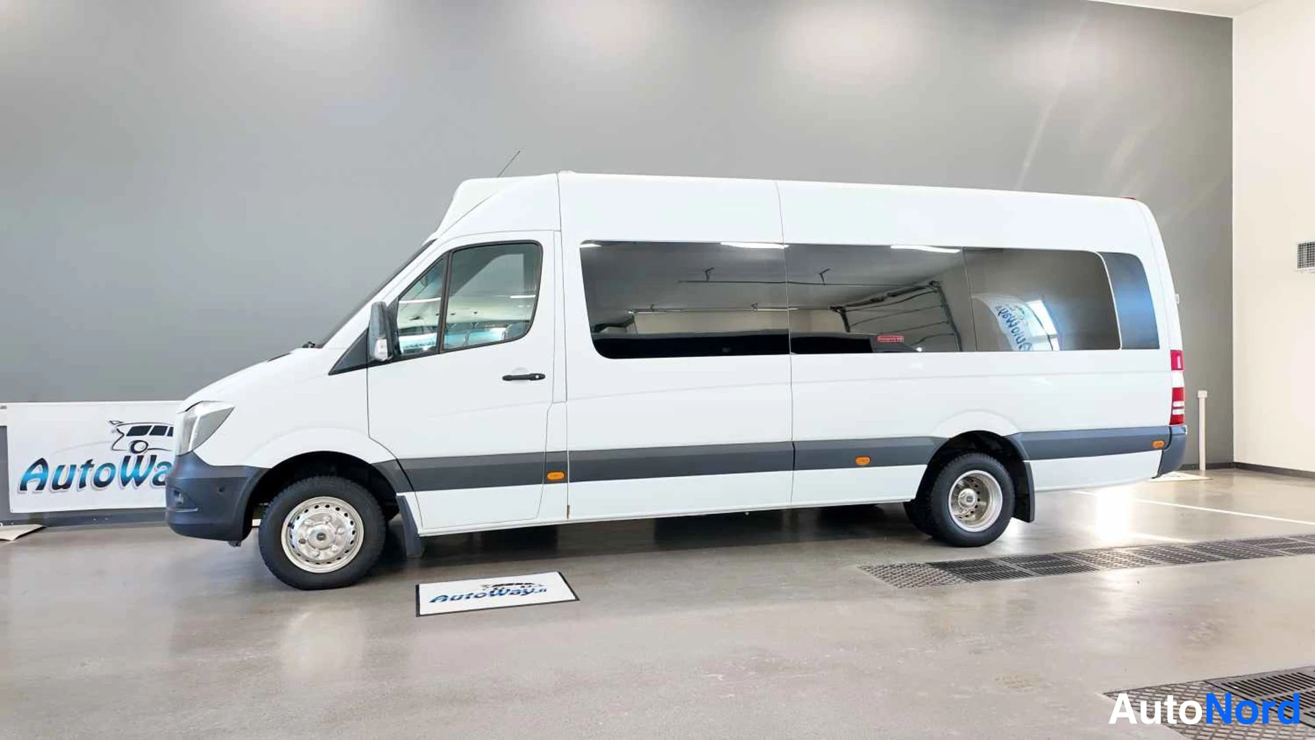 Sivulta: Mercedes-Benz Sprinter -pituussuuntainen pakettibussi valkoisena sivuprofiilissa hallin lattialla, pitkä matkustamo ja iso takasivuikkuna.