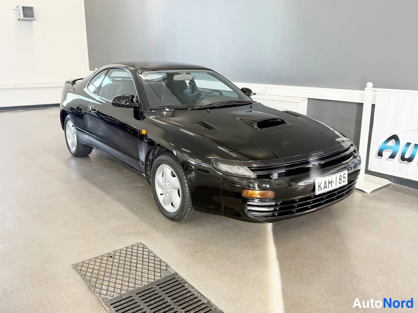 Etuviisto, nokka oikealle: Mustan Toyota Celican etukulmakolmasosa sisätiloissa, rekisterikilpi näkyvissä.