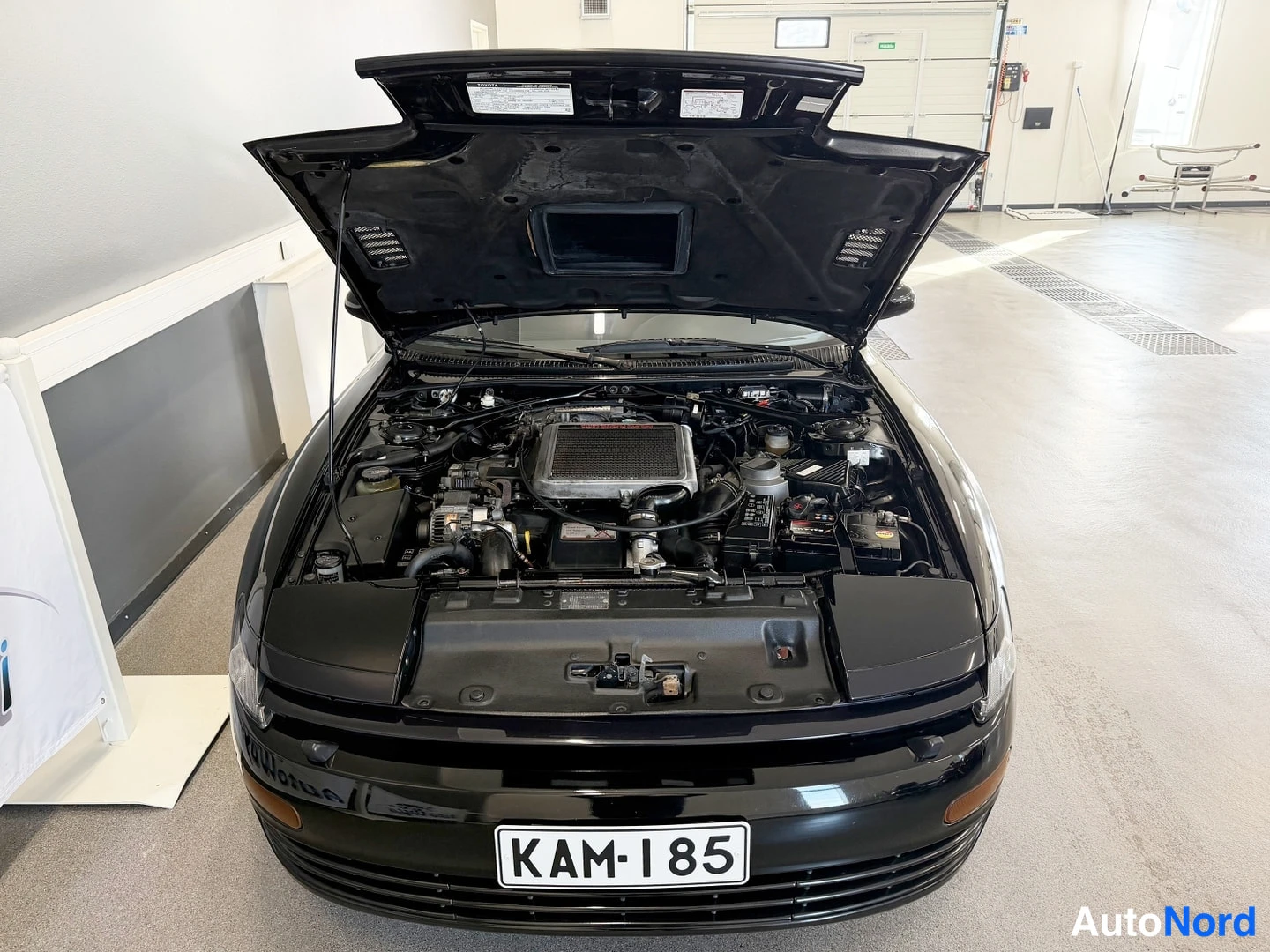Edestä: Musta Toyota Celica konepelti auki, moottoritila näkyvissä rekisterikilpi.