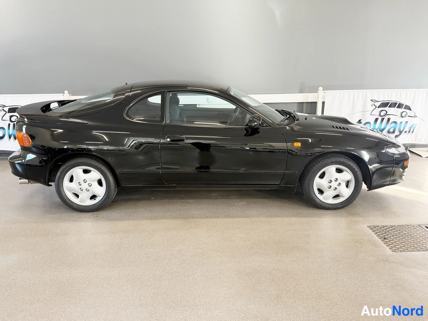 Sivulta: Mustan Toyota Celican sivuprofiili sisätiloissa, takasiipi ja takaosa näkyvät.
