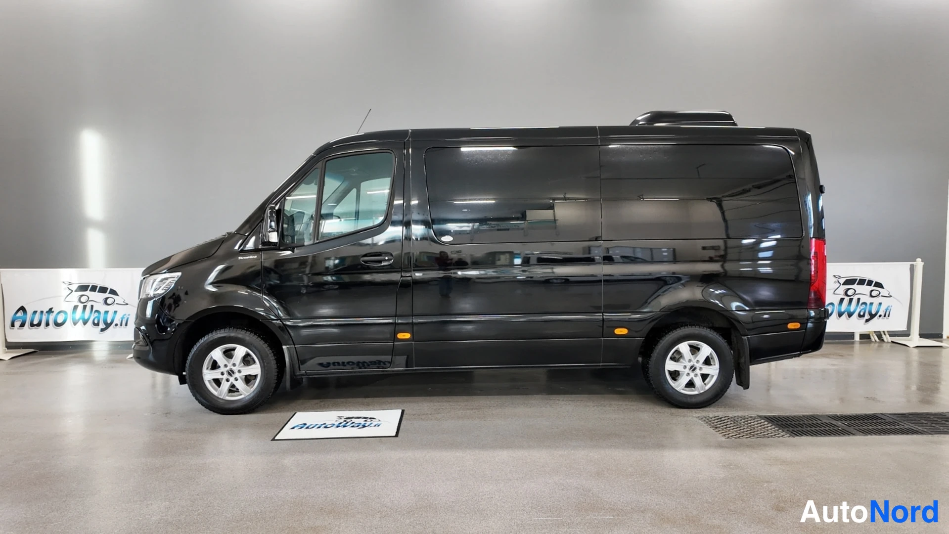 Sivulta: Musta Mercedes-Benz Sprinter sivulta vasemmalta, koko kylki hallissa näkyvissä.
