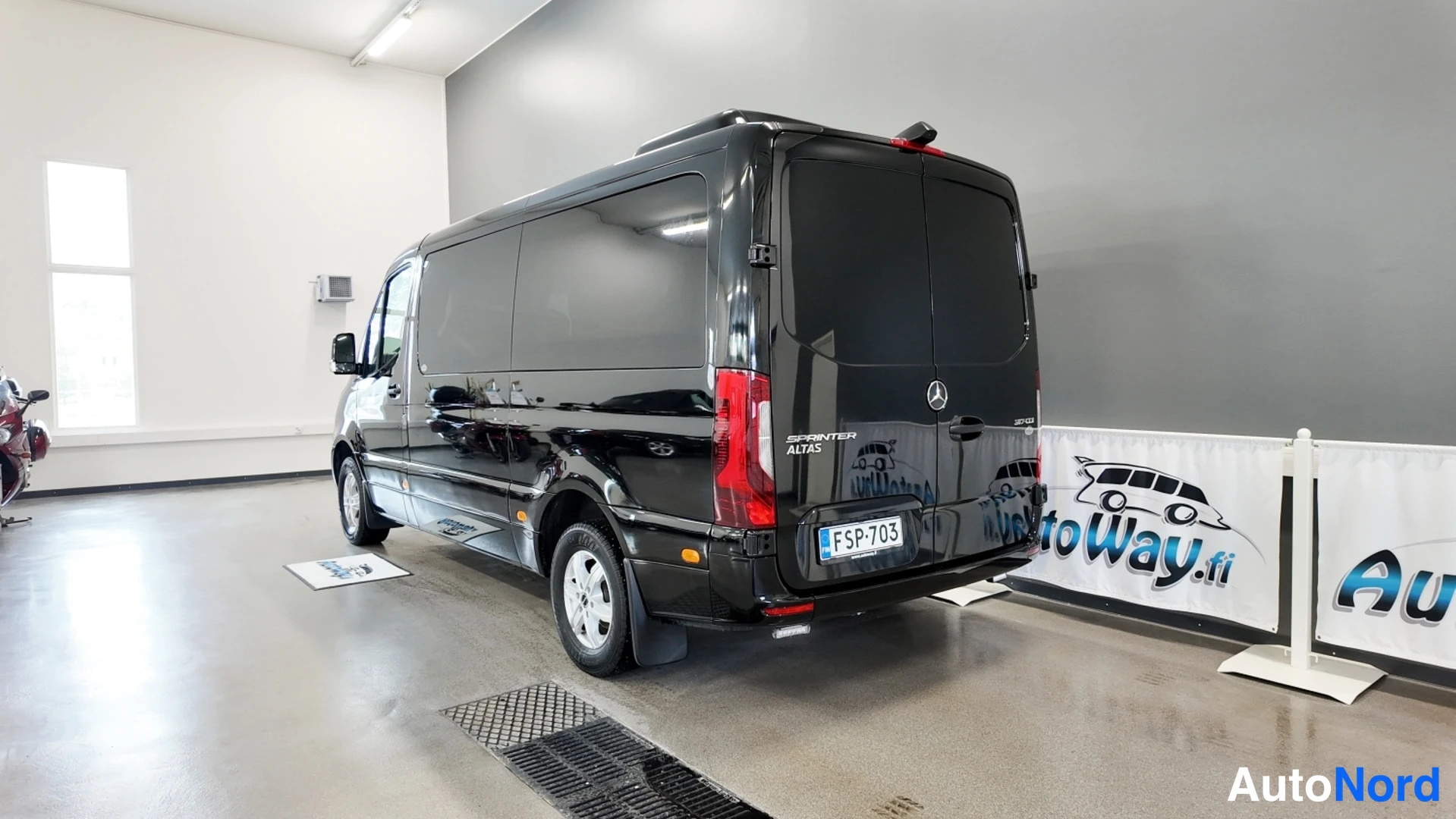 Takaviisto: Mustan Mercedes-Benz Sprinterin takakulma vasemmalta, takapuoli ja kylki hallissa näkyvissä.