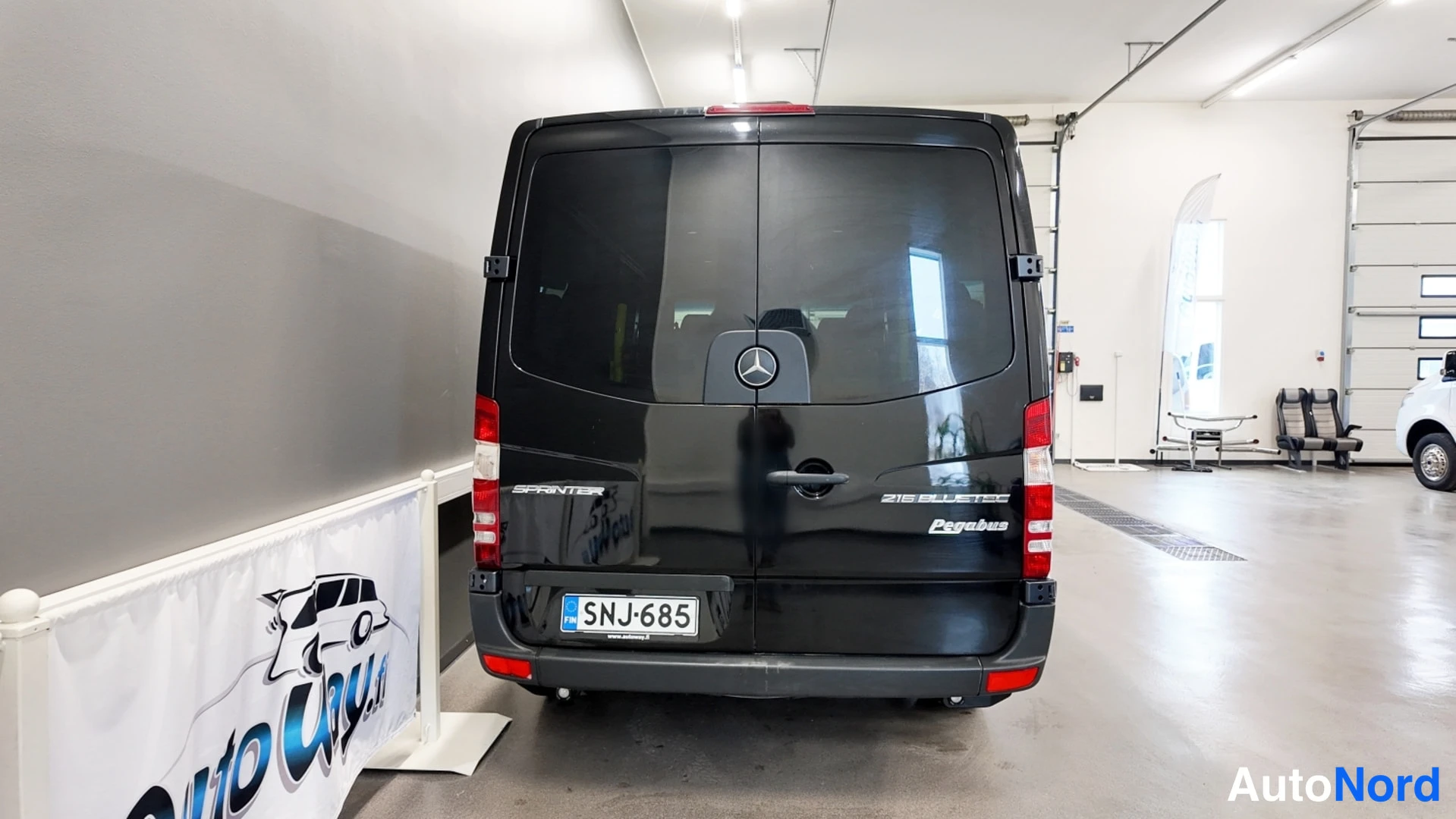Takaa: Mustan pakettiauton takanäky suoraan takaa hallissa, rekisterikilpi.