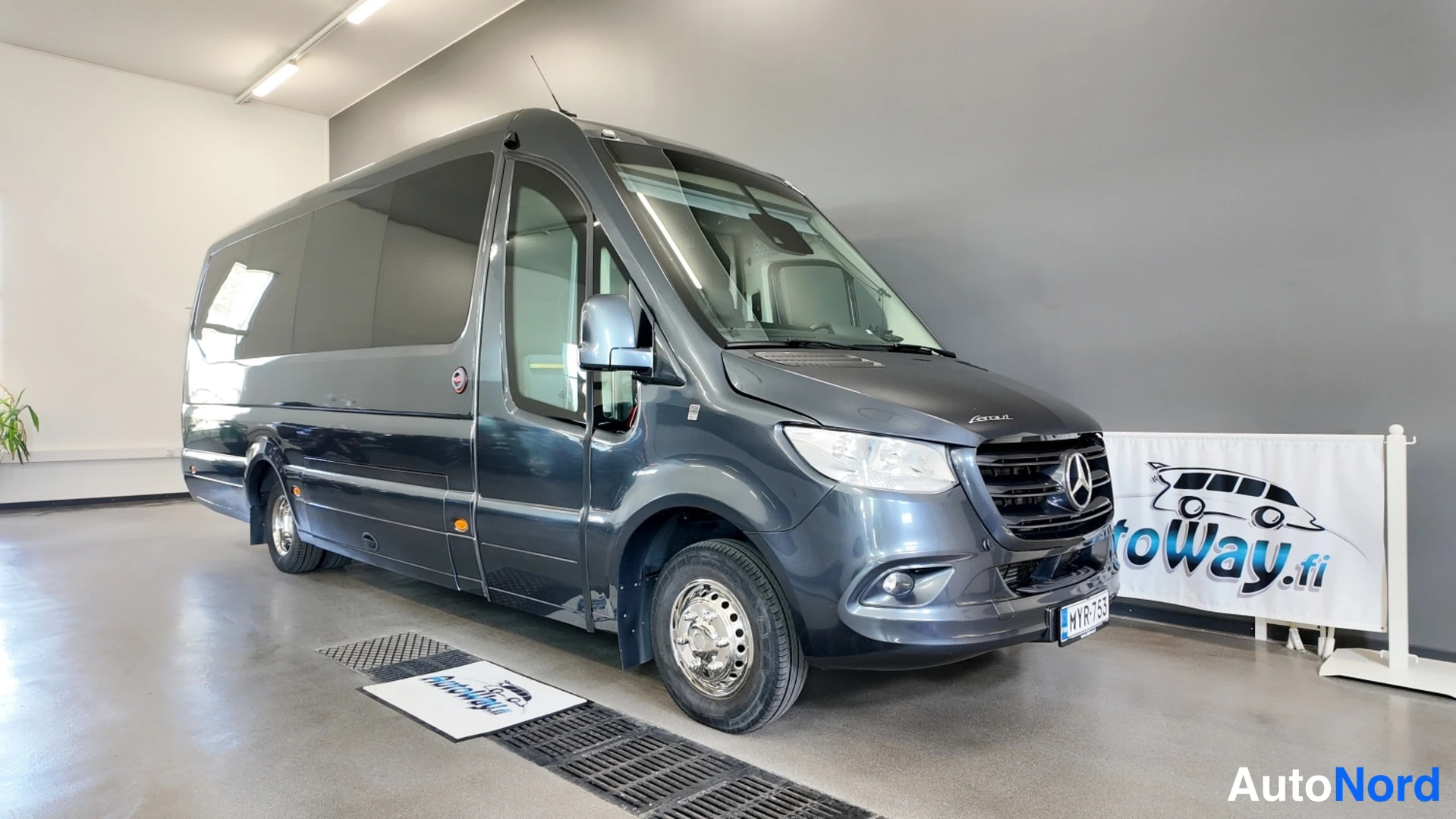Etuviisto, nokka oikealle: Tummanharmaa Mercedes-Benz Sprinter -bussi kulmassa etuosa ja sivu näkyvissä, koriin avattu sivuovi ja kylkeen asennetut ikkunat.