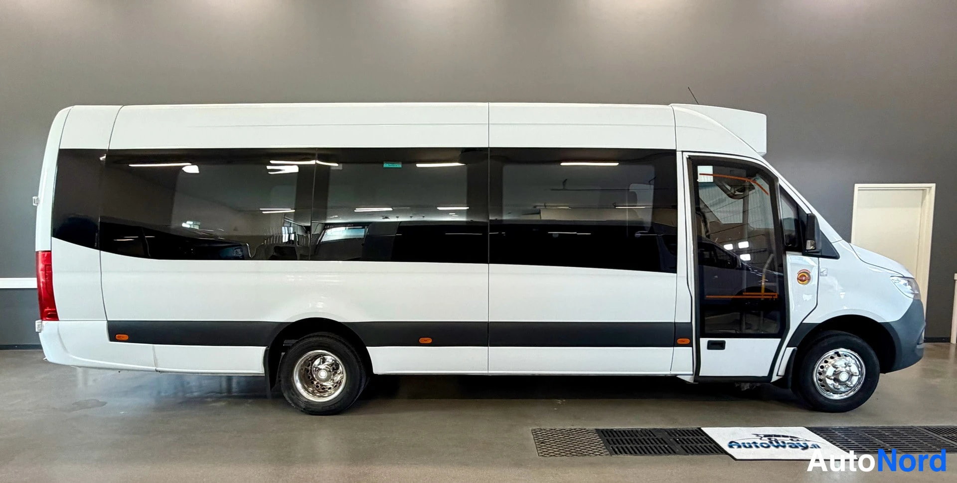 Sivulta: Mercedes-Sprinter-pohjaisen linja-autokorin valkoinen sivuprofiili, pitkät tummat ikkunat, sivuovi auki-asennossa, kuva sisätilassa.