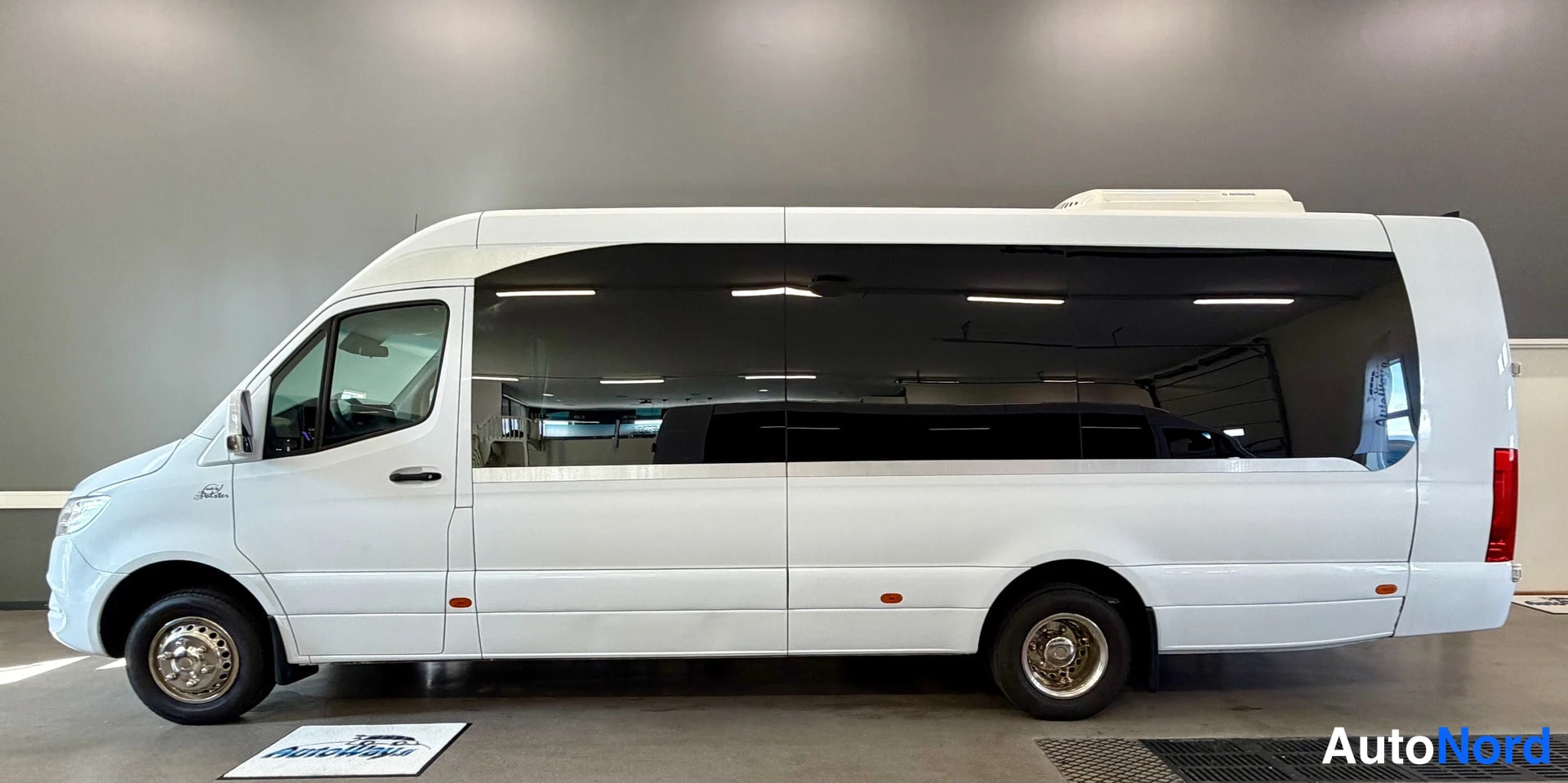 Sivulta: Mercedes-Benz Sprinter -pakettiauton sivuprofiili; valkoinen korimusta ikkunalinja ja takapyörät näkyvät hallissa.