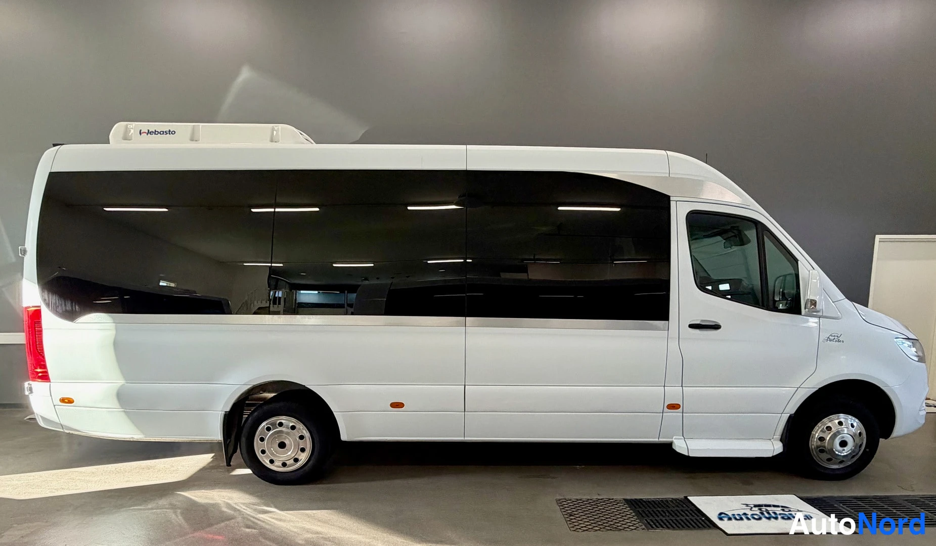Sivulta: Valkoisen Mercedes-Benz Sprinter -bussin sivunäkymä sisätiloissa, pitkä sivuseinä ikkunoineen.