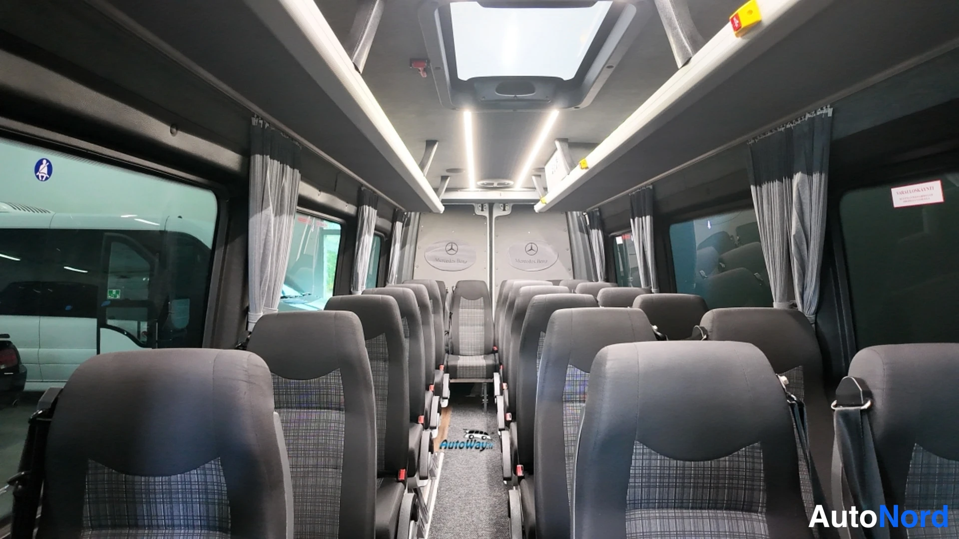 Muu: Pakettiautobussin sisätilat takaapäin: kaksi harmaiden verhoiltujen matkustajistuolien rivistä, kattokaiuttimet, LED-valot, kattoluukku ja sivuikkunoissa verhot