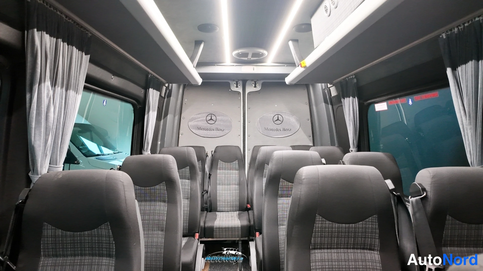 Muu: Pakettiautobussin matkustamo takaa päin kuvattuna: harmaat verhoillut istuimet rivissä, välikäytävä, kattoluukku ja Mercedes-tähti takaovessa.