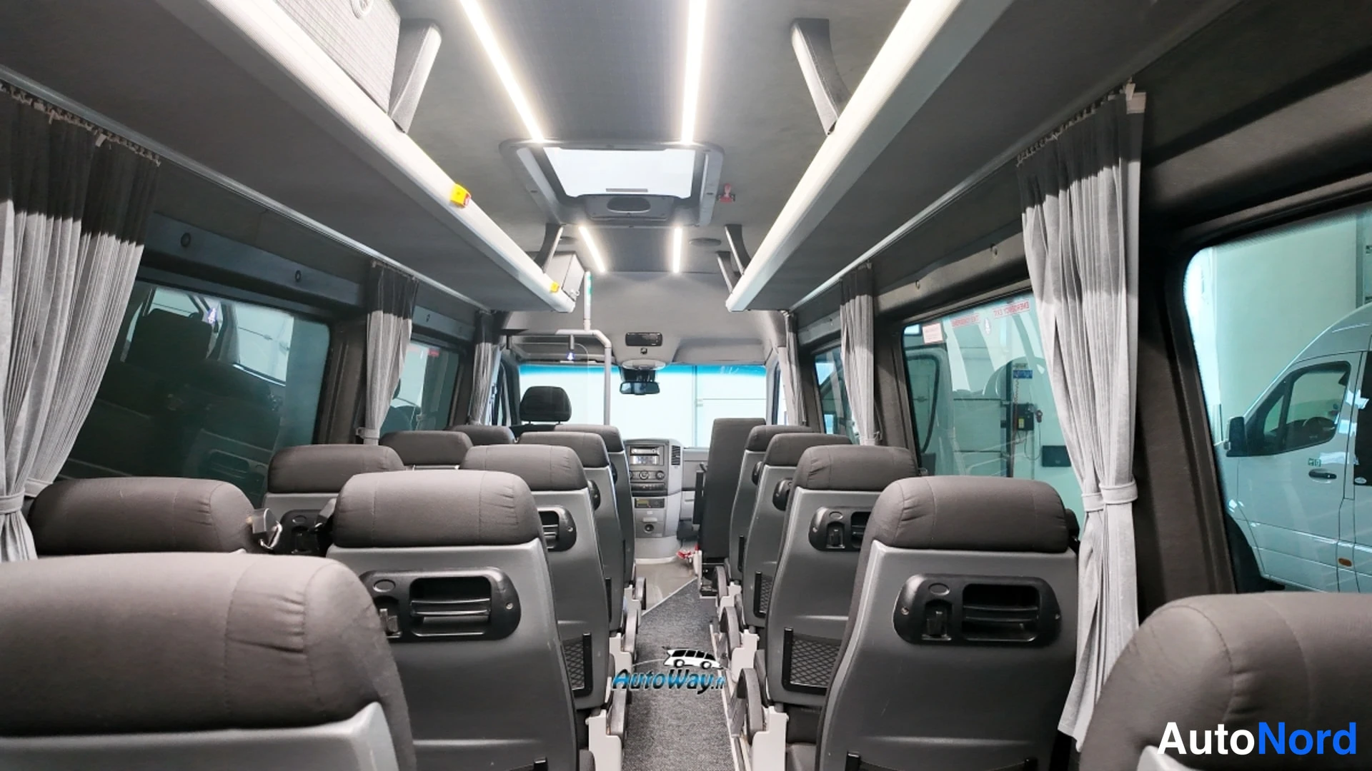 Muu: Pakettiautobussin sisätila keulaa kohti: harmaat matkustajistuolien rivit, kattoluukku, LED-kattovalot ja valkoiset verhot sivuikkunoissa.