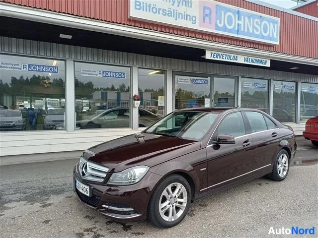 Etuviisto, nokka vasemmalle: Tummanpunainen Mercedes-Benz C-sedan paikallaan autoliikkeen R Johnsonin edustalla, keulakulma vasemmalle.