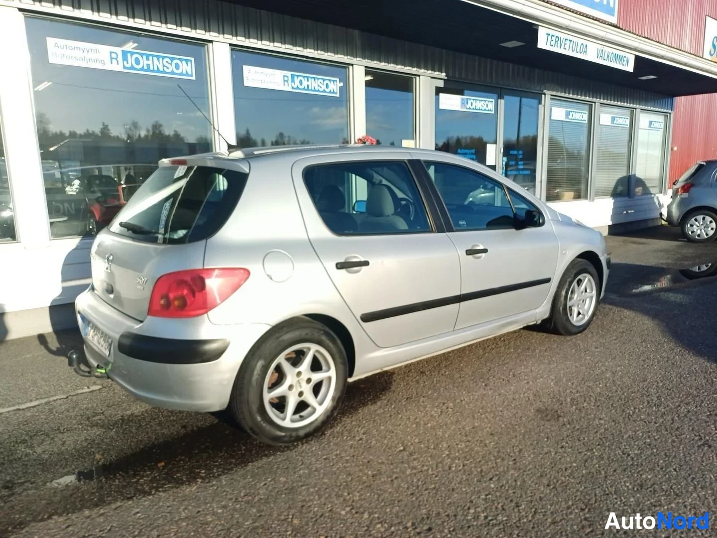 Takaviisto: Hopeanvärinen Peugeot 307 viistosti takaa-autotallin ikkunoita vasten, vasen kylki ja takapää näkyvissä.