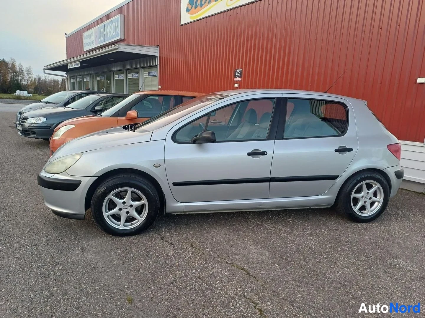 Sivulta: Hopeanvärinen viisiovinen Peugeot 307 -mallin sivunäkymä edestä vasemmalle kuvattuna punaisen hallirakennuksen edessä.