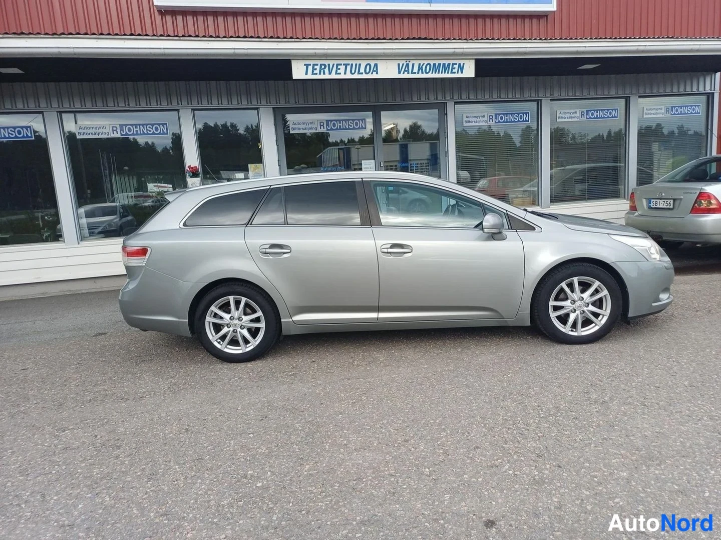 Sivulta: Hopeanharmaan Toyota Avensis -farmarin sivuprofiili, autonliikkeen ikkunoiden edessä.