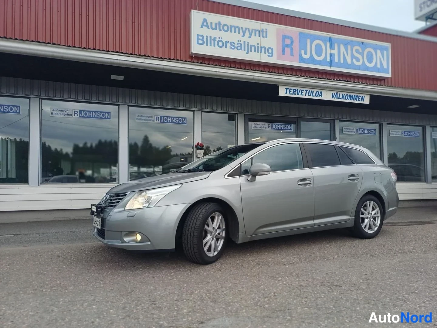Etuviisto, nokka vasemmalle: Toyota Avensis -farmarin etu-kulmanäkymä; hopea kori, sumuvalot päällä, edessä Automyynti R Johnsonin liiketalo