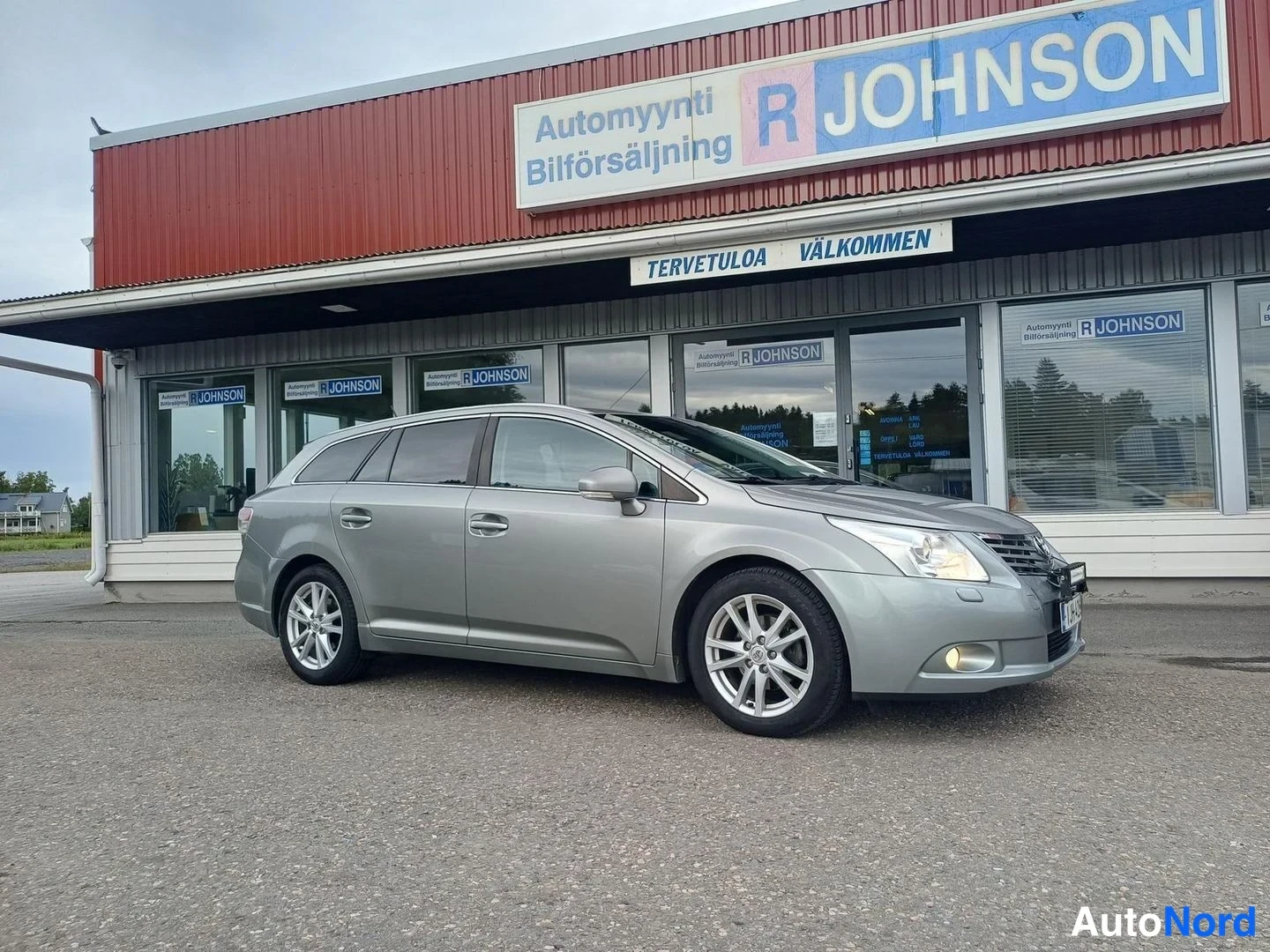 Etuviisto, nokka oikealle: Hopeanharmaa Toyota Avensis -farmari etu kolmasosakulmasta, nokka oikealla, autonliikkeen edessä.
