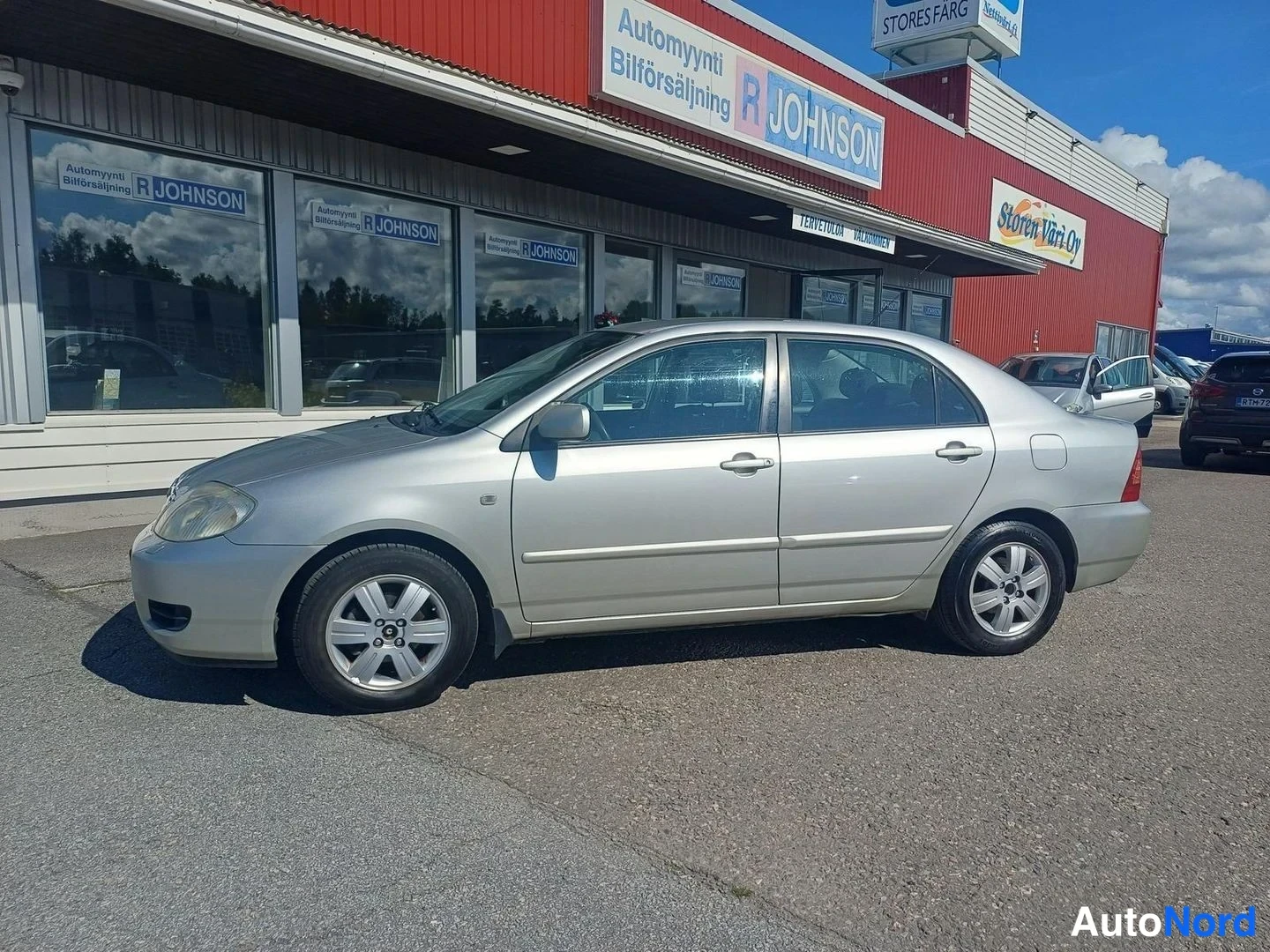 Etuviisto, nokka vasemmalle: Toyota Corolla -sedanin etukulma vasemmalta sivulta, hopeinen kori liikkeen ikkunoiden edessä.