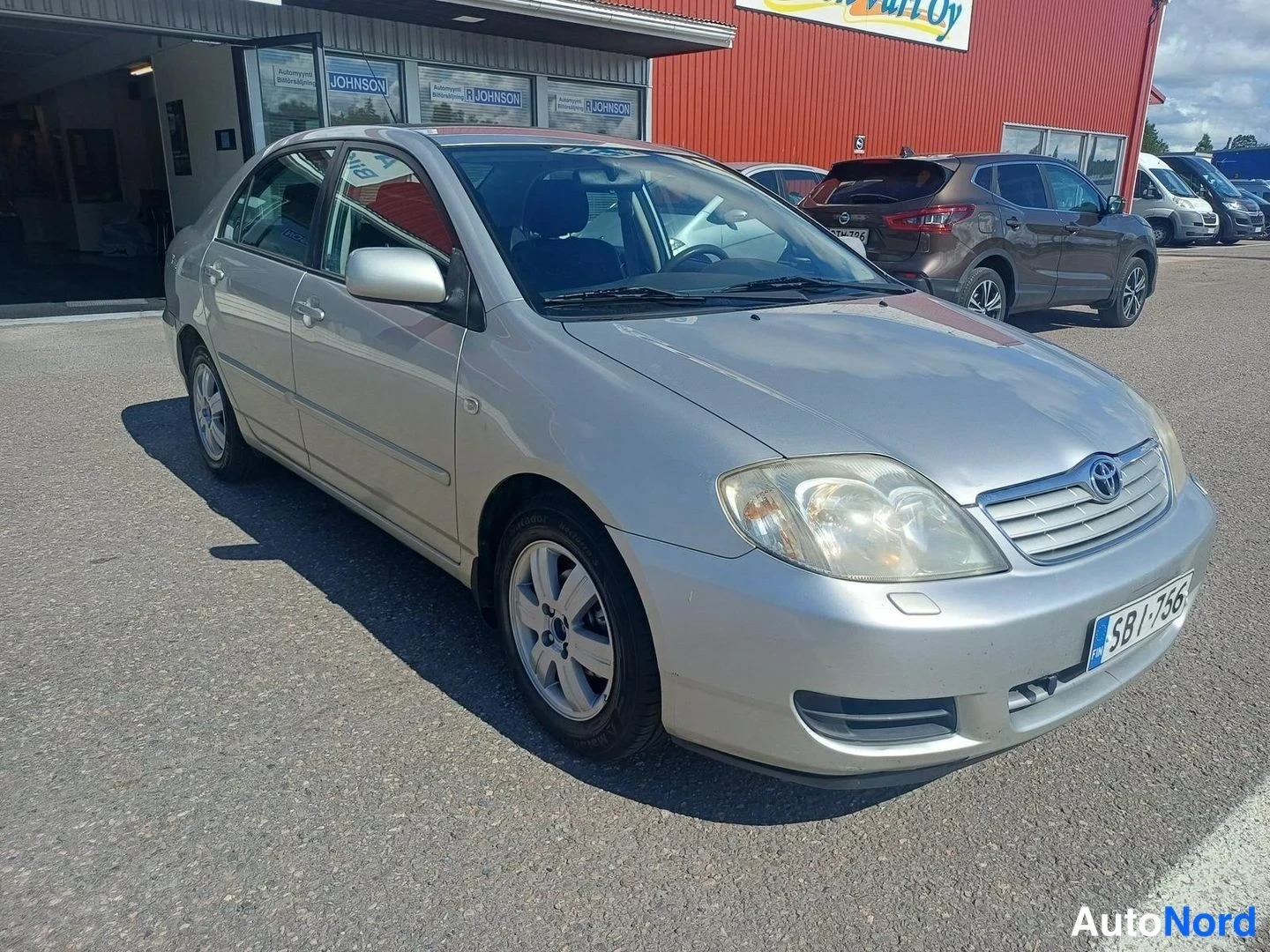 Etuviisto, nokka oikealle: Toyota Corolla -sedanin etukulma oikealta, hopeinen kori parkkipaikalla, rekisteritunnus.