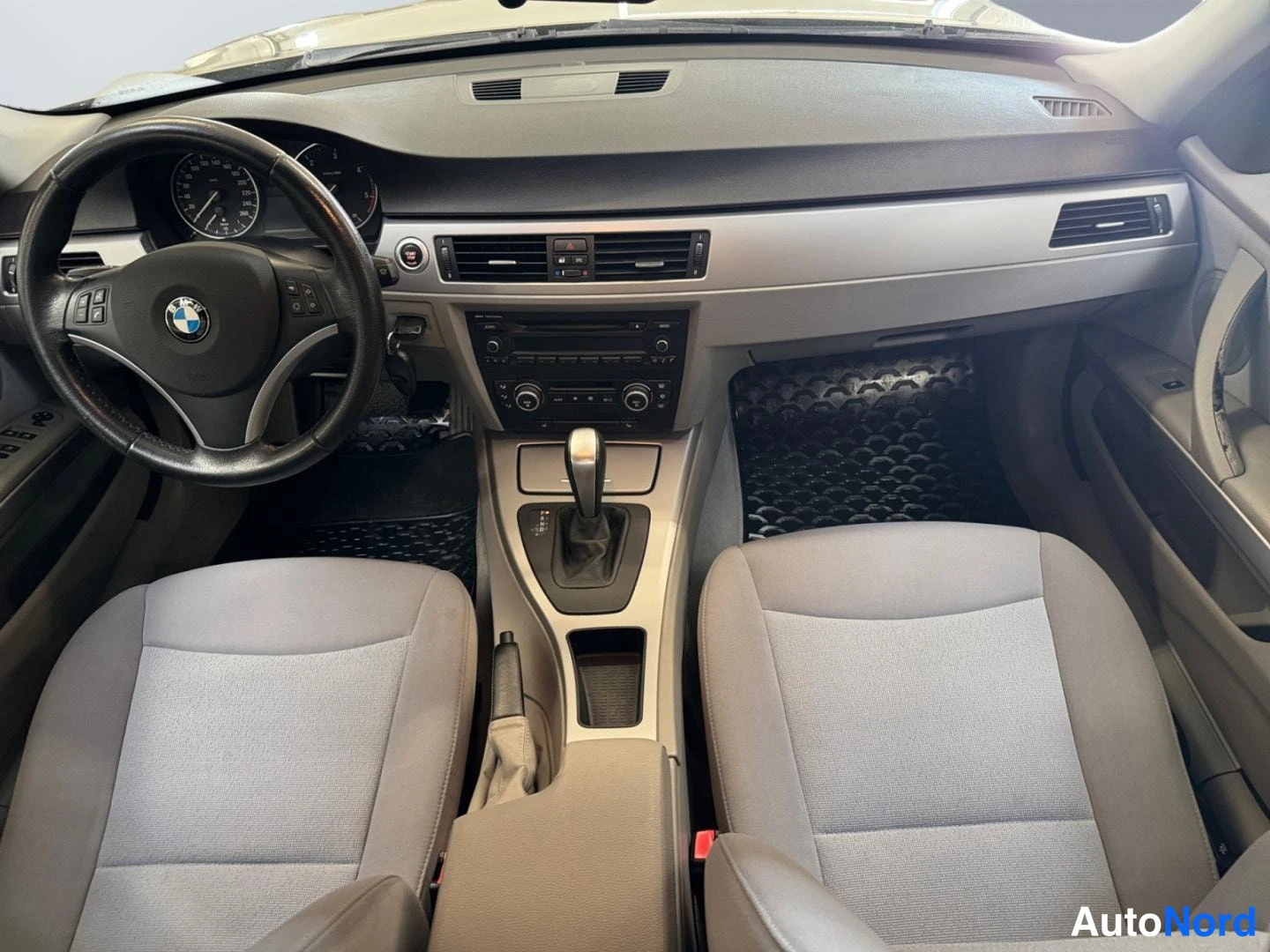 Etuviisto: Etusivu: ohjauspyörä BMW-merkillä, analogiset mittarit, automaattivalitsin ja keskikonsolin radiopaneeli; vaaleat kankaatpenkit ja mustat matot.