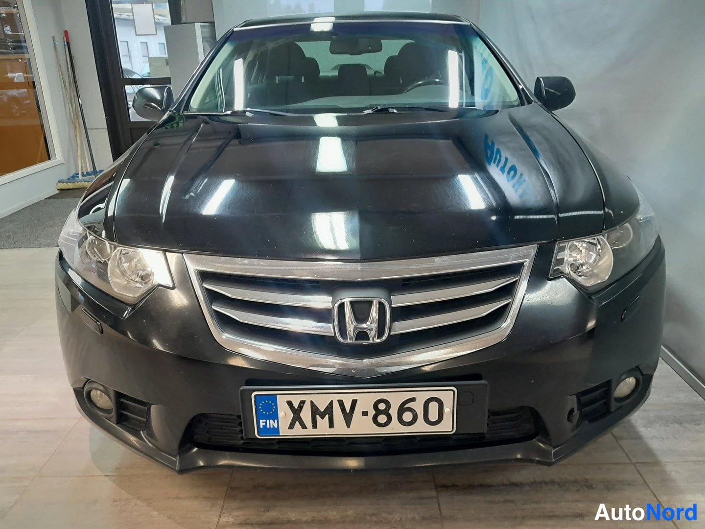 Edestä: Mustan Honda Accord -sedanin keula näkyvissä: etumootti, H-merkki, kiiltävät parkit ja suomalainen rekisterikilpi sisätiloissa.
