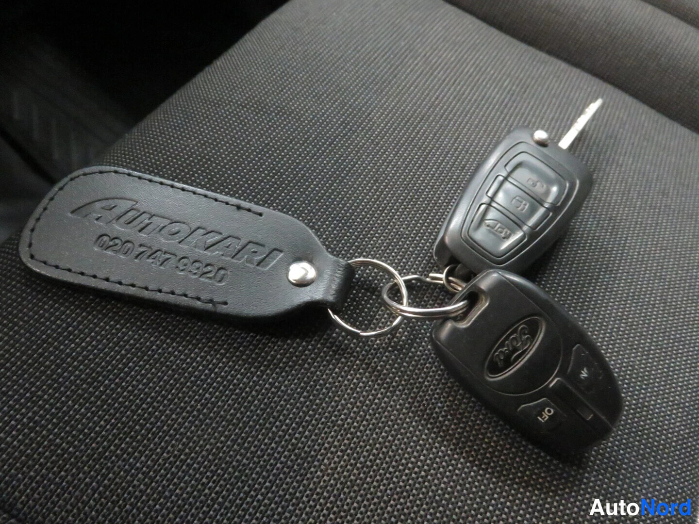 Muu: Avaimenperä ja kaksi auton avain laitteen istuinkankaan päällä; perässä lukee Autokari 020 747 9920.