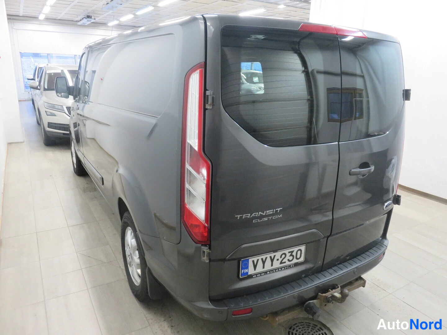 Takaviisto: Ford Transit Custom -pakettiauton takaosan ja vasemman sivun näkymä sisätiloissa, rekisterikilpi ja TRANSIT CUSTOM -teksti takapaneelissa; autonväri tummanharma