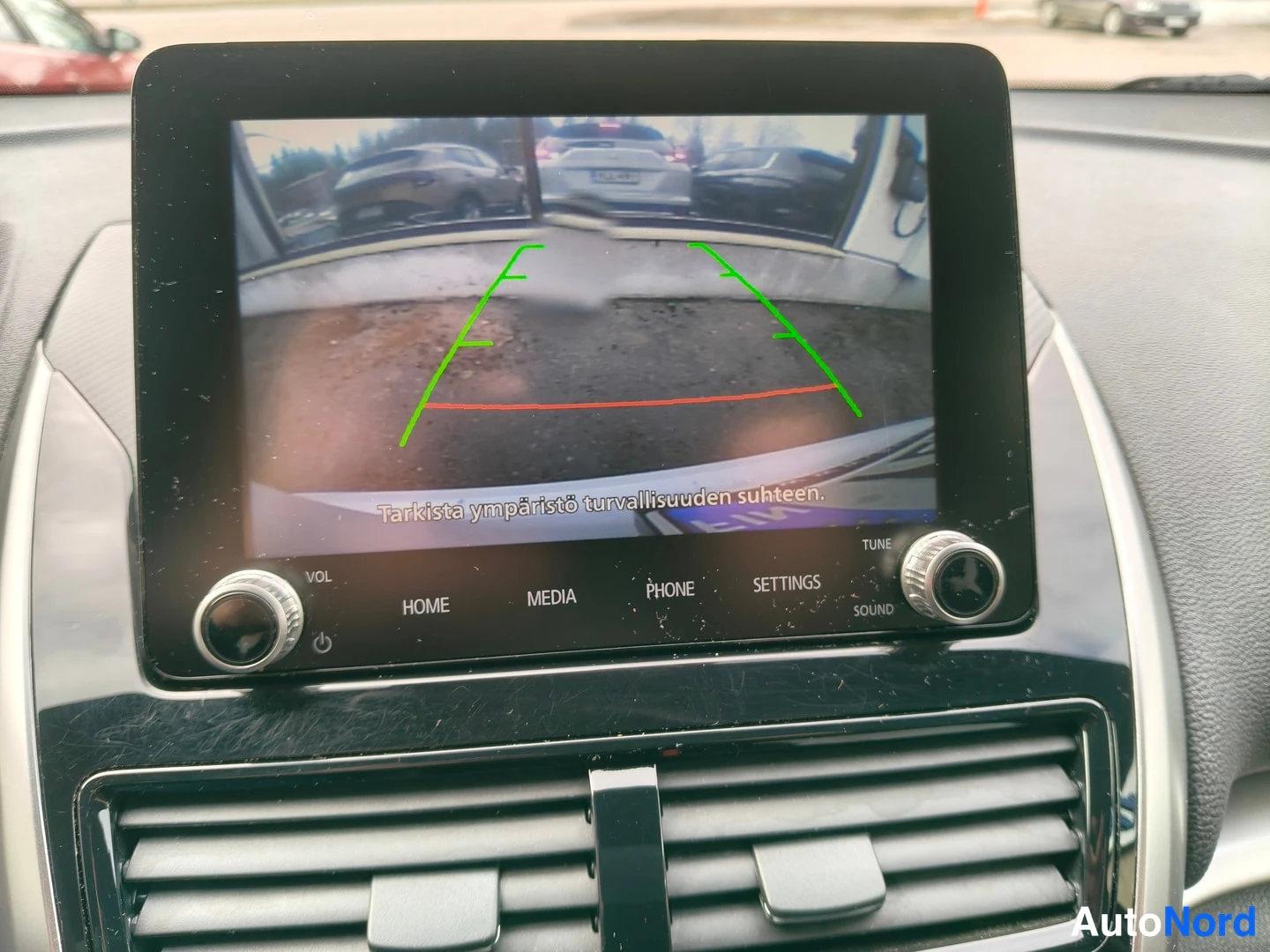 Etuviisto, nokka vasemmalle: Ylösnouseva infotainment-näyttö näyttää peruutuskameran kuvan ohjauslinjoineen ja tekstillä 'Tarkista ympäristö turvallisuuden suhteen.'