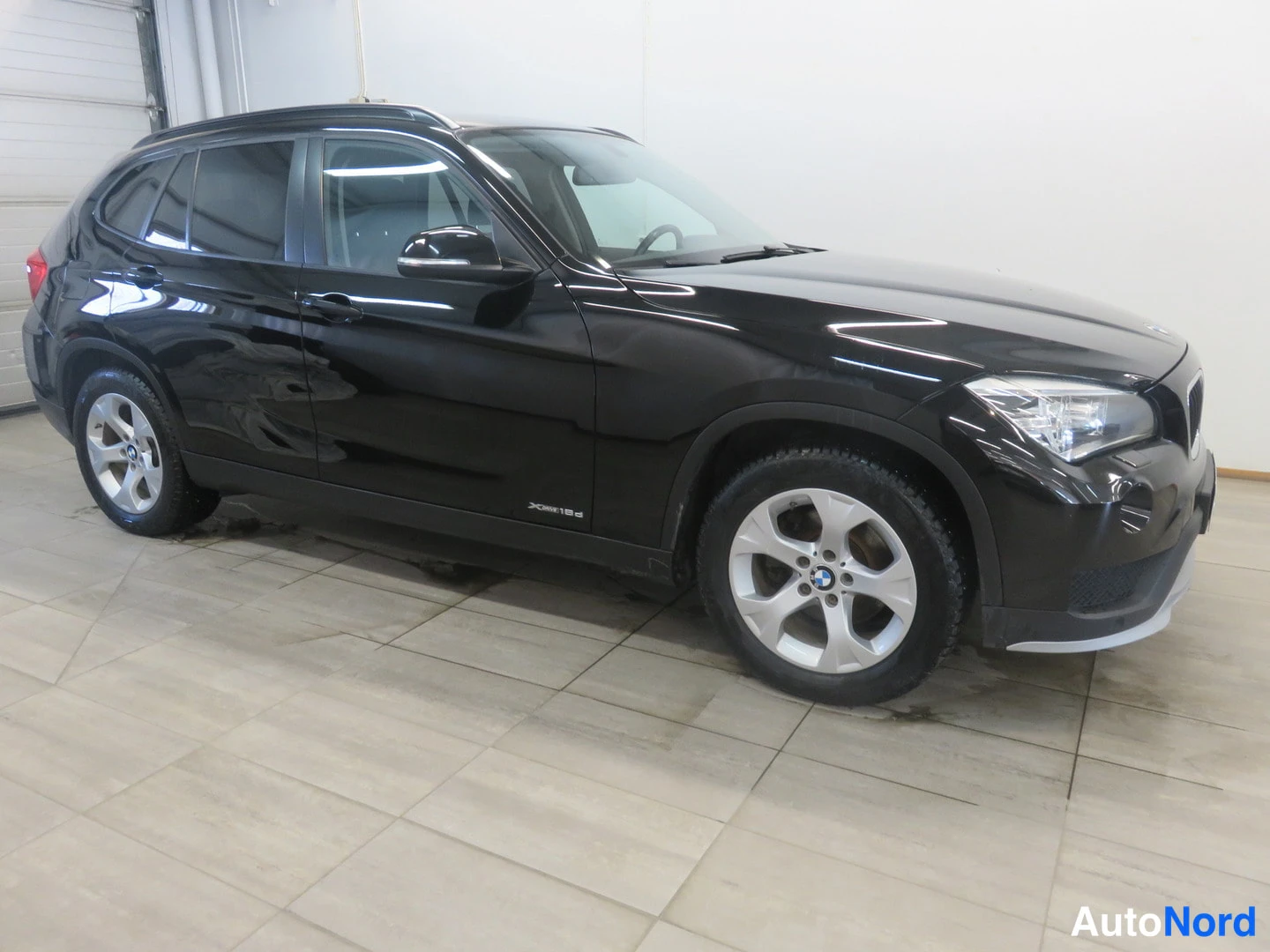 Etuviisto, nokka oikealle: Musta BMW X1 -maastopoika kulmassa oikealta etupäältä kuvattuna sisätilassa.