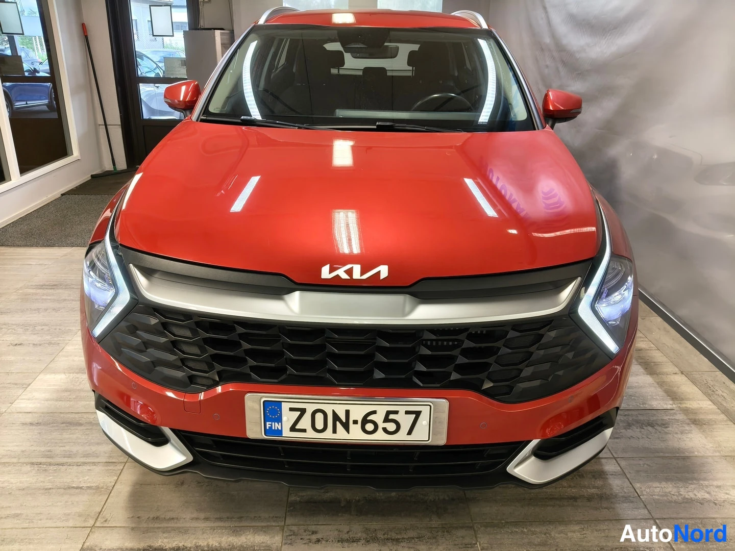 Edestä: Punaisen KIA Sportage -maasturin keula suoraan edestä, KIA-merkki etusäleikössä ja rekisteritunnus.