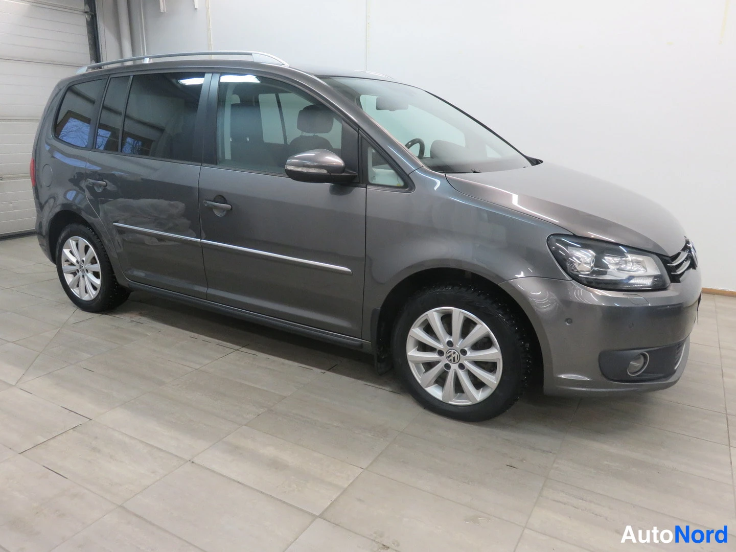 Etuviisto, nokka oikealle: Harmaan Volkswagen Touran -pakettiauton etukulma sisätiloissa, kuljettajan puoli näkyvissä.