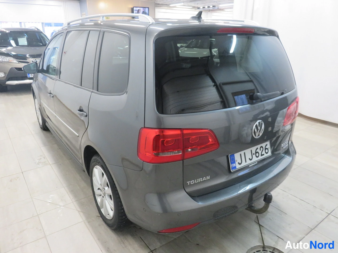 Takaviisto: Auton takakulma: tummanharmaan Volkswagen Touranin takaosa ja vasen kylki näkyvät sisätiloissa, rekisteritunnus.